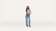 A woman stands against a plain white background, wearing a light floral blouse with puffed shoulders, tucked into high-waisted wide-leg blue jeans and open-toe shoes. She has one hand in her pocket and looks forward.