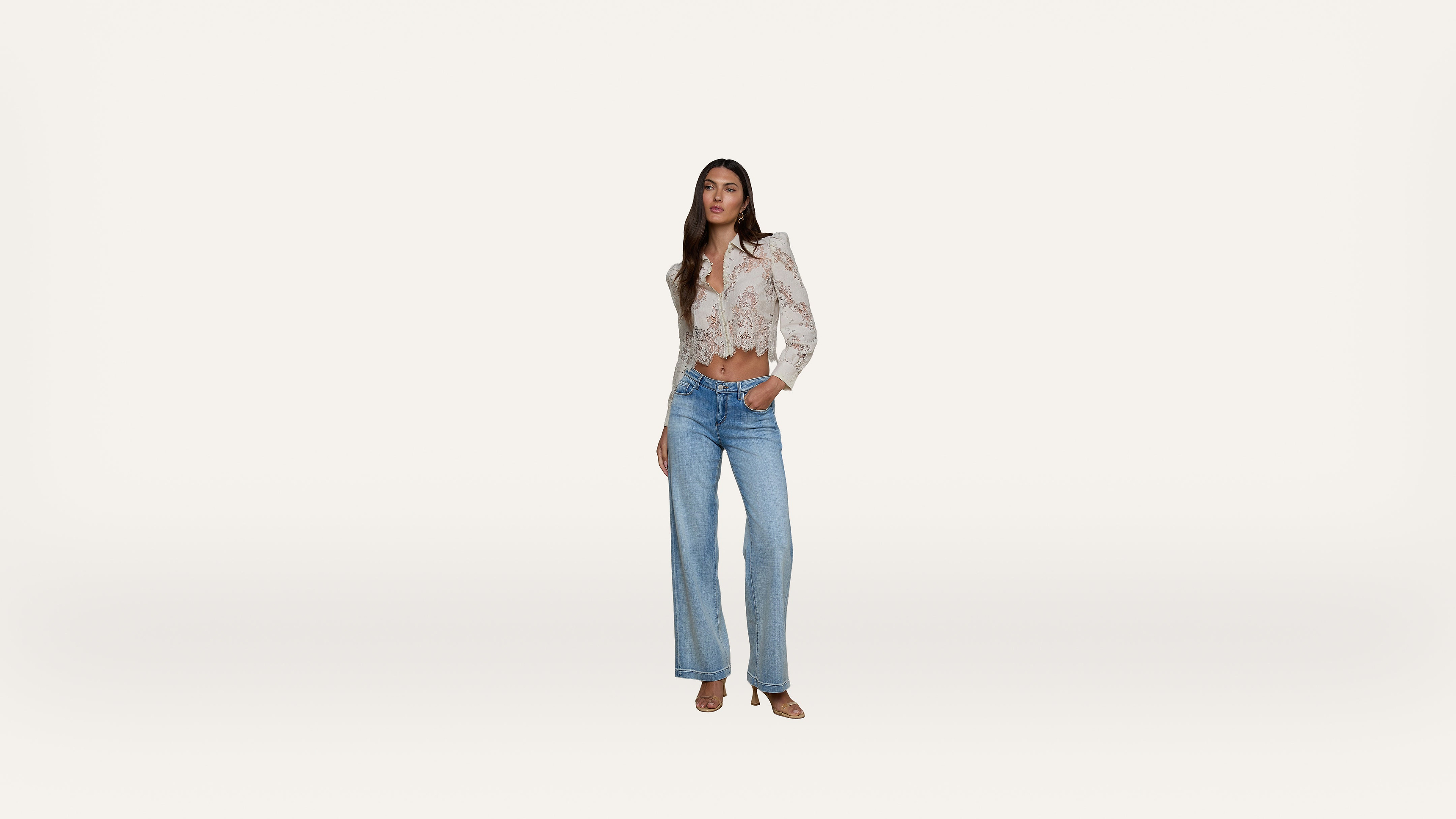 A woman stands against a plain white background, wearing a light floral blouse with puffed shoulders, tucked into high-waisted wide-leg blue jeans and open-toe shoes. She has one hand in her pocket and looks forward.