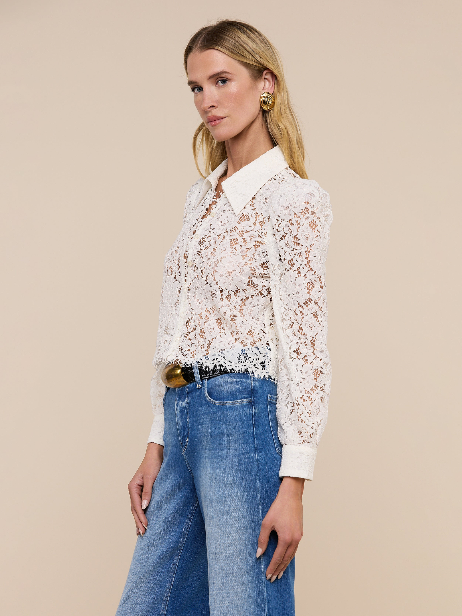 Wearing the Anita Lace Blouse—a romantic sheer white floral lace blouse with a collar and long sleeves—paired with blue high-waisted jeans and gold earrings, a woman stands against a plain light background.