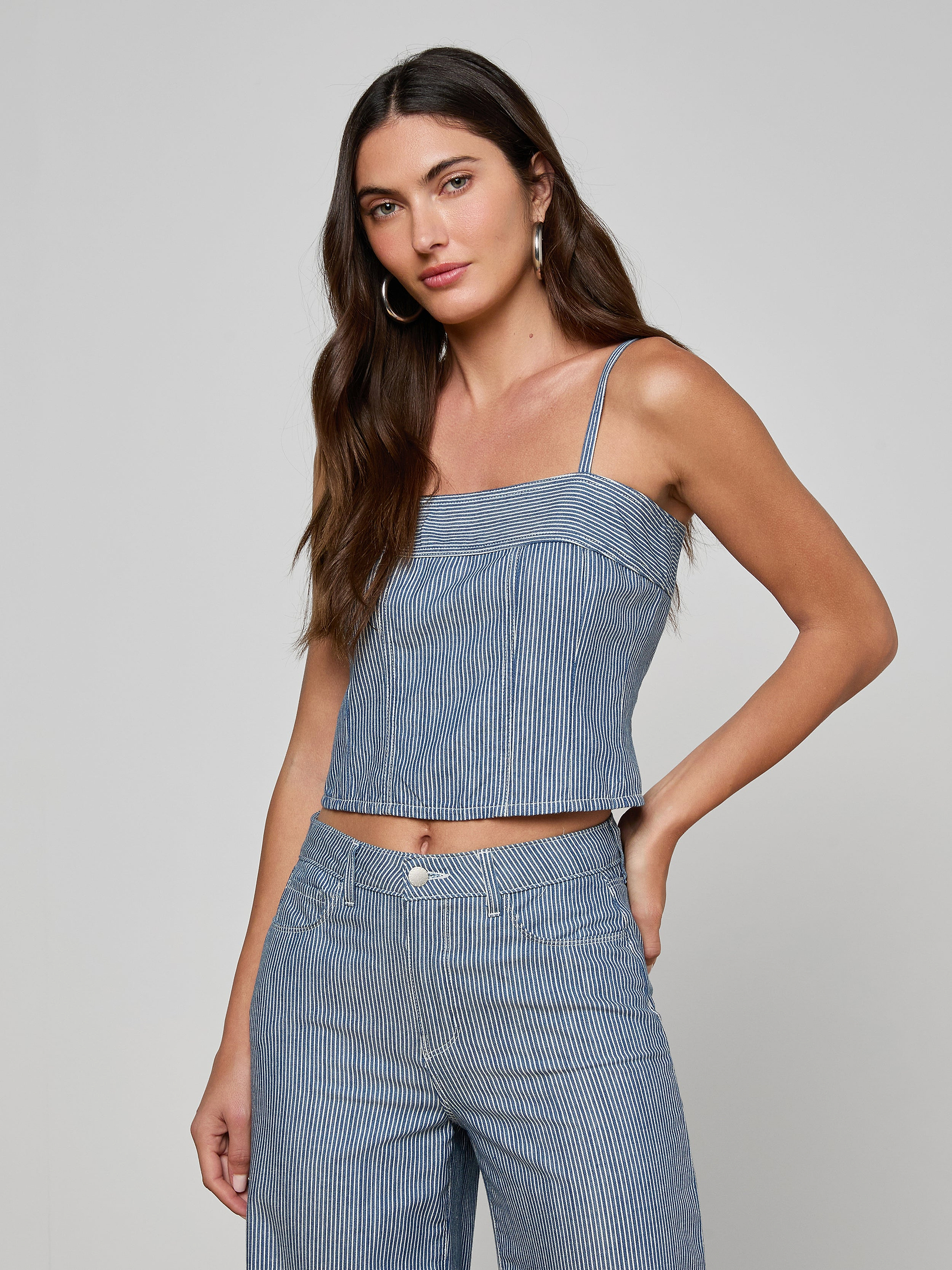 A woman with long brown hair is wearing the Arlin Tank—a blue striped sleeveless top—paired with matching pants, standing with one hand on her hip against a plain light background.