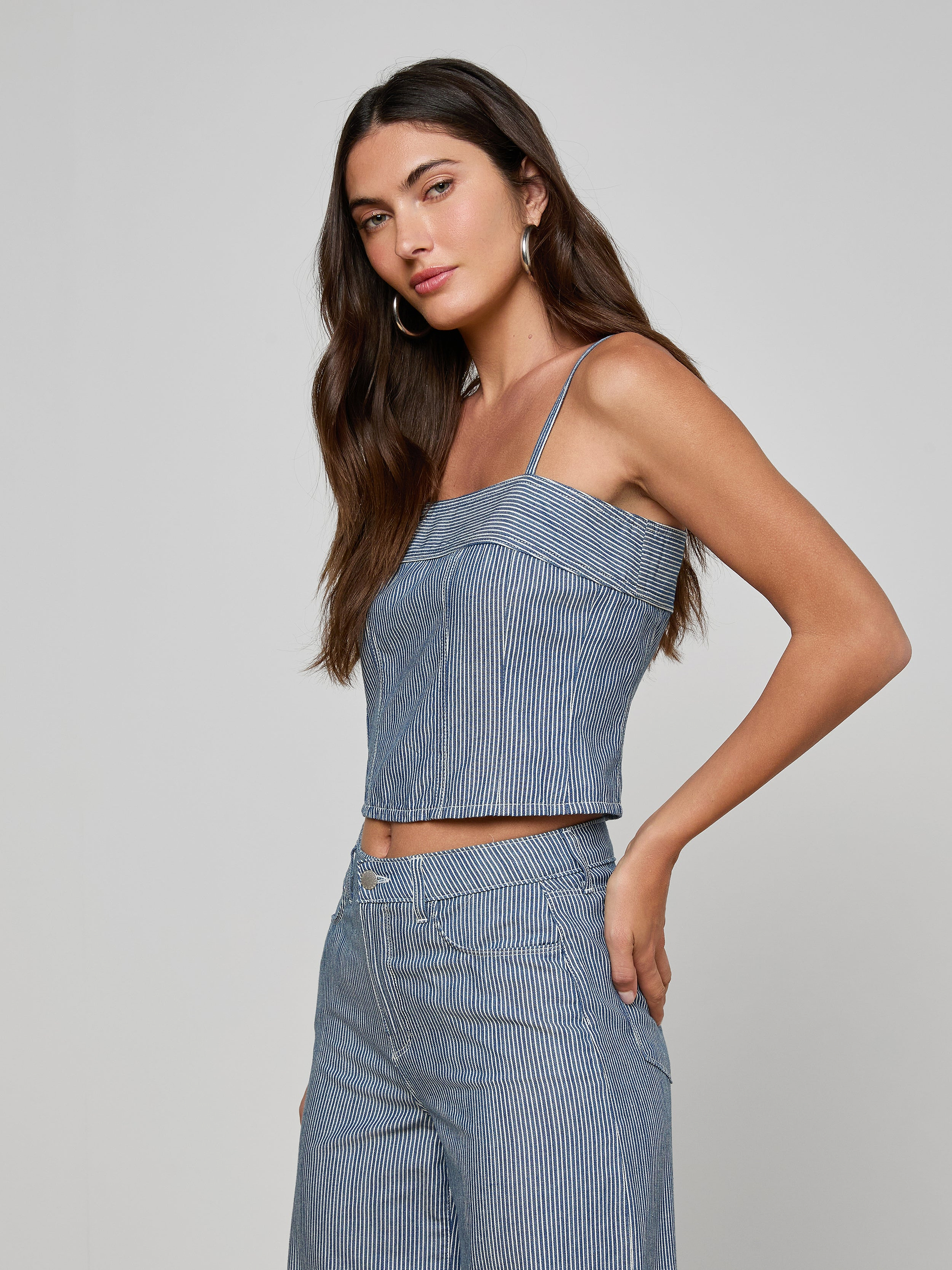 Woman wearing the Arlin Tank, a blue stripe sleeveless corset top paired with matching high-waisted pants, standing against a plain light background and looking at the camera with one hand on her hip.