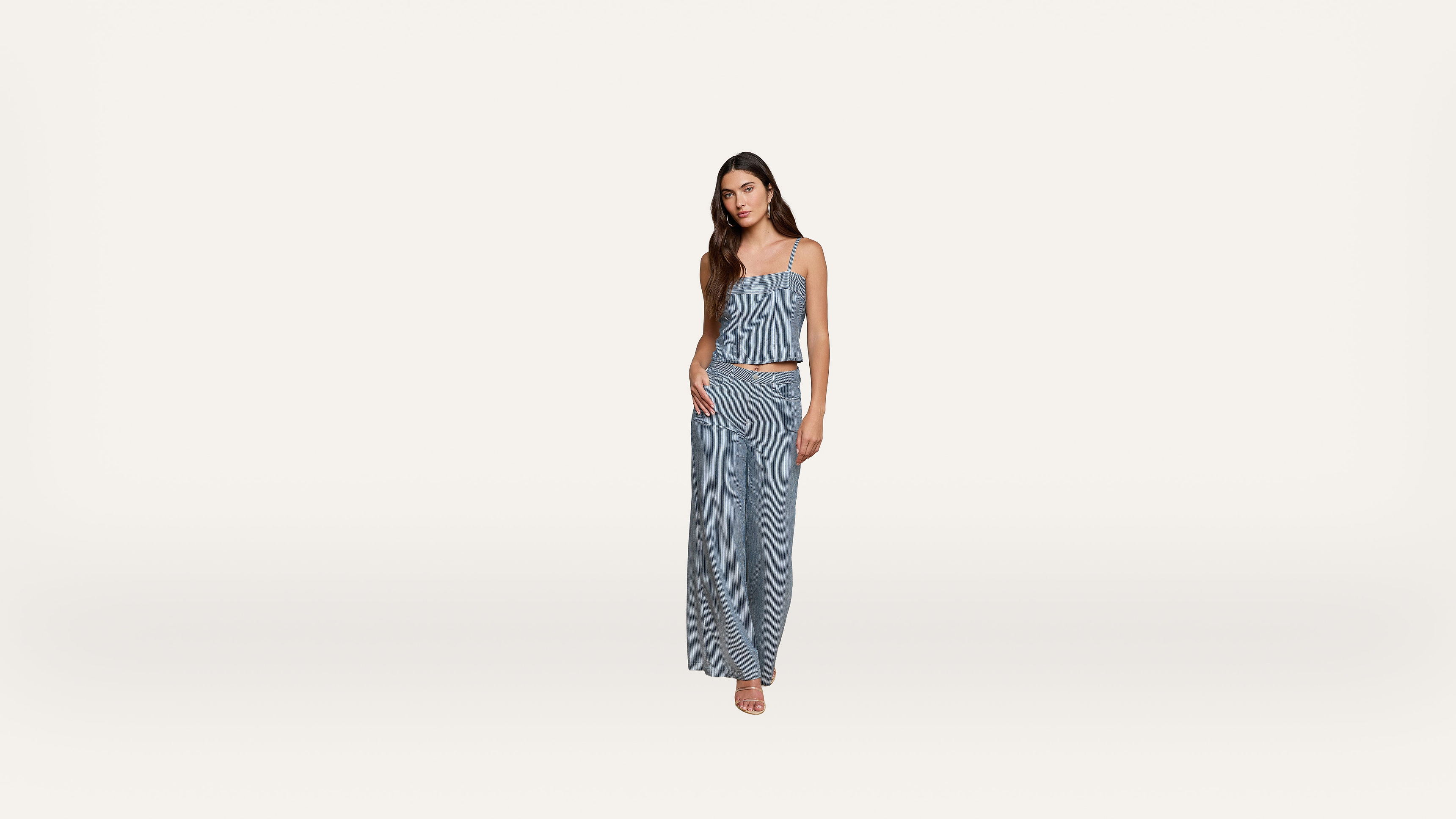 A woman stands against a plain white background, wearing a blue strapless top and matching wide-leg pants. She has long dark hair and neutral sandals, with one hand resting on her hip.