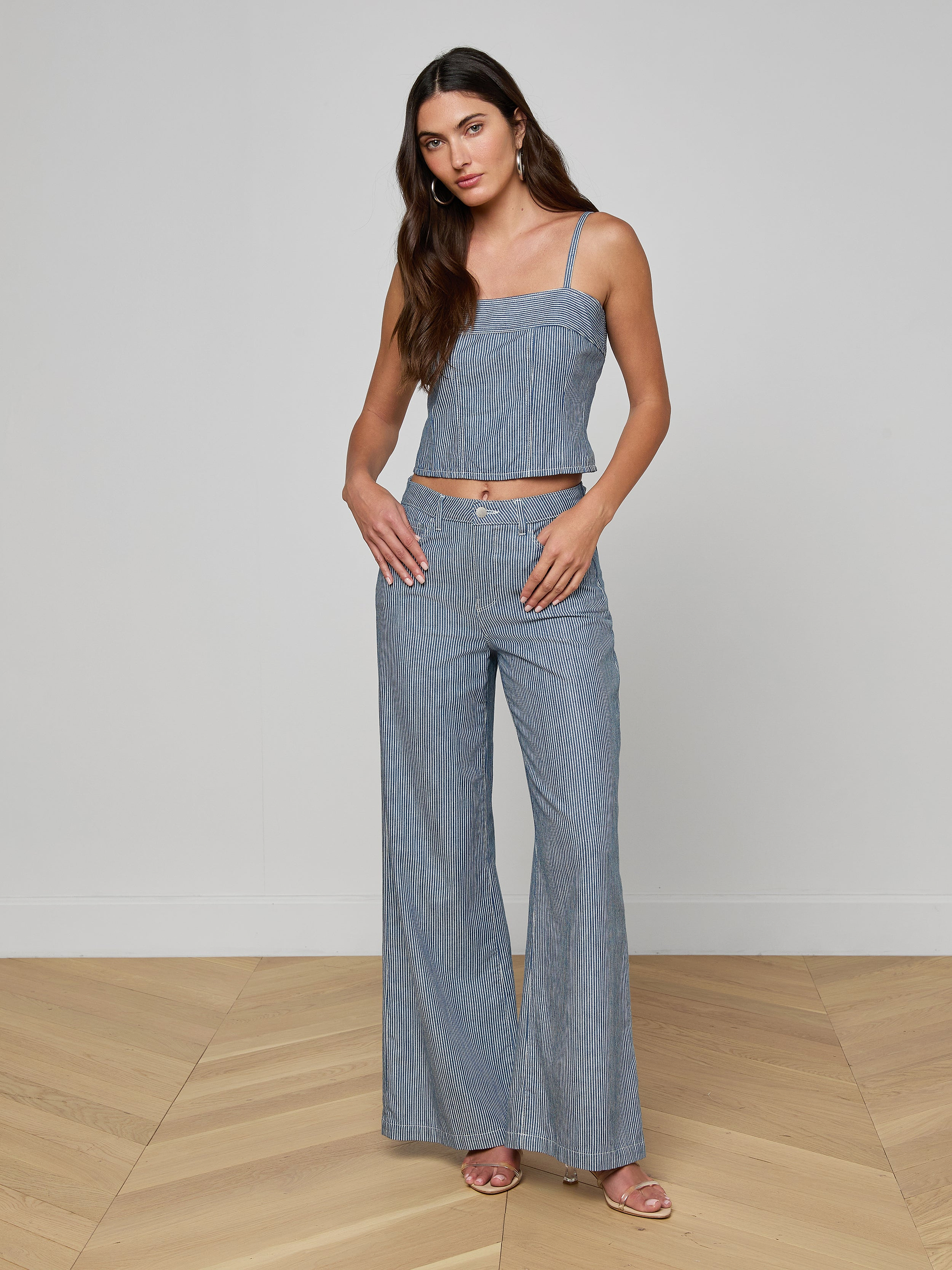 A woman stands against a plain background, wearing the Arlin Tank—a sleeveless blue stripe corset-style top—paired with matching high-waisted, wide-leg pants. Her long hair is down and she looks at the camera with her hands in her pockets.
