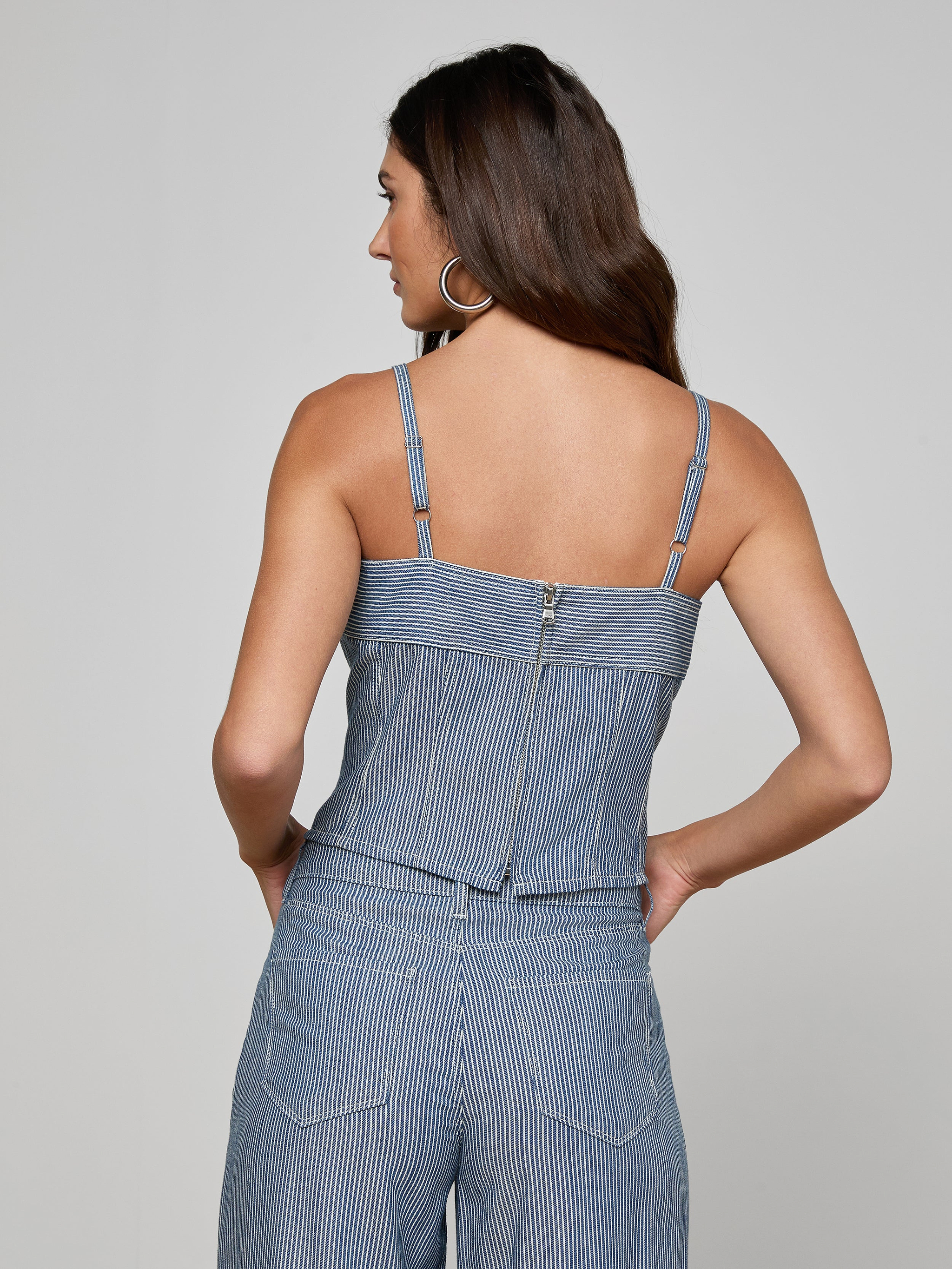Woman facing away, wearing the Arlin Tank—a blue pinstriped sleeveless top with thin straps, a visible back zipper, and subtle corset-inspired detailing—paired with matching pinstriped pants. Her hands are in her pockets and she has long brown hair.