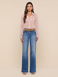 A woman stands against a plain white background, wearing the Laurent Blouse—a light pink, sheer poly chiffon button-up shirt with long sleeves and a relaxed fit—paired with blue high-waisted wide-leg jeans, a brown belt, and open-toe shoes.
