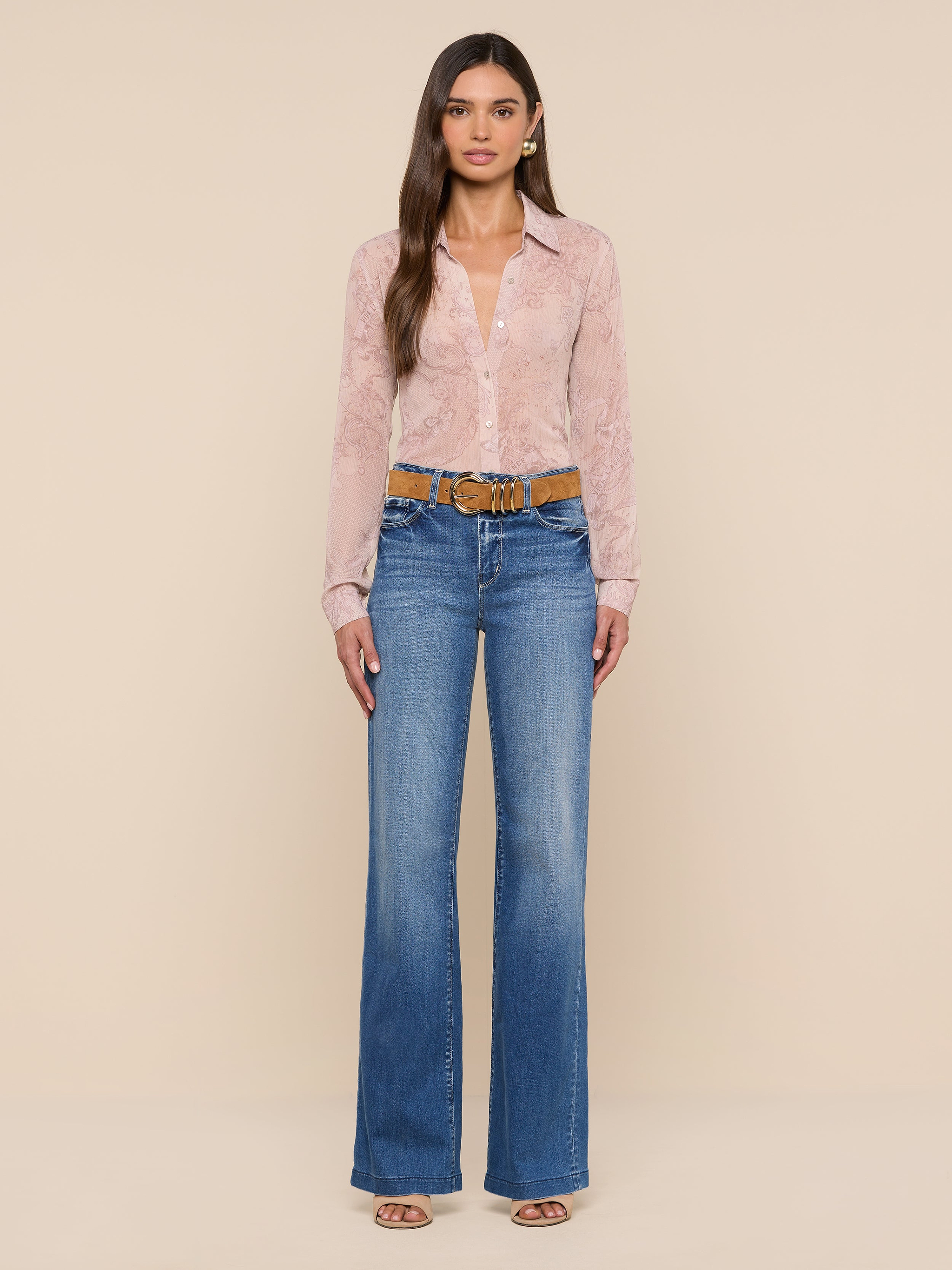 A woman stands against a plain white background, wearing the Laurent Blouse—a light pink, sheer poly chiffon button-up shirt with long sleeves and a relaxed fit—paired with blue high-waisted wide-leg jeans, a brown belt, and open-toe shoes.