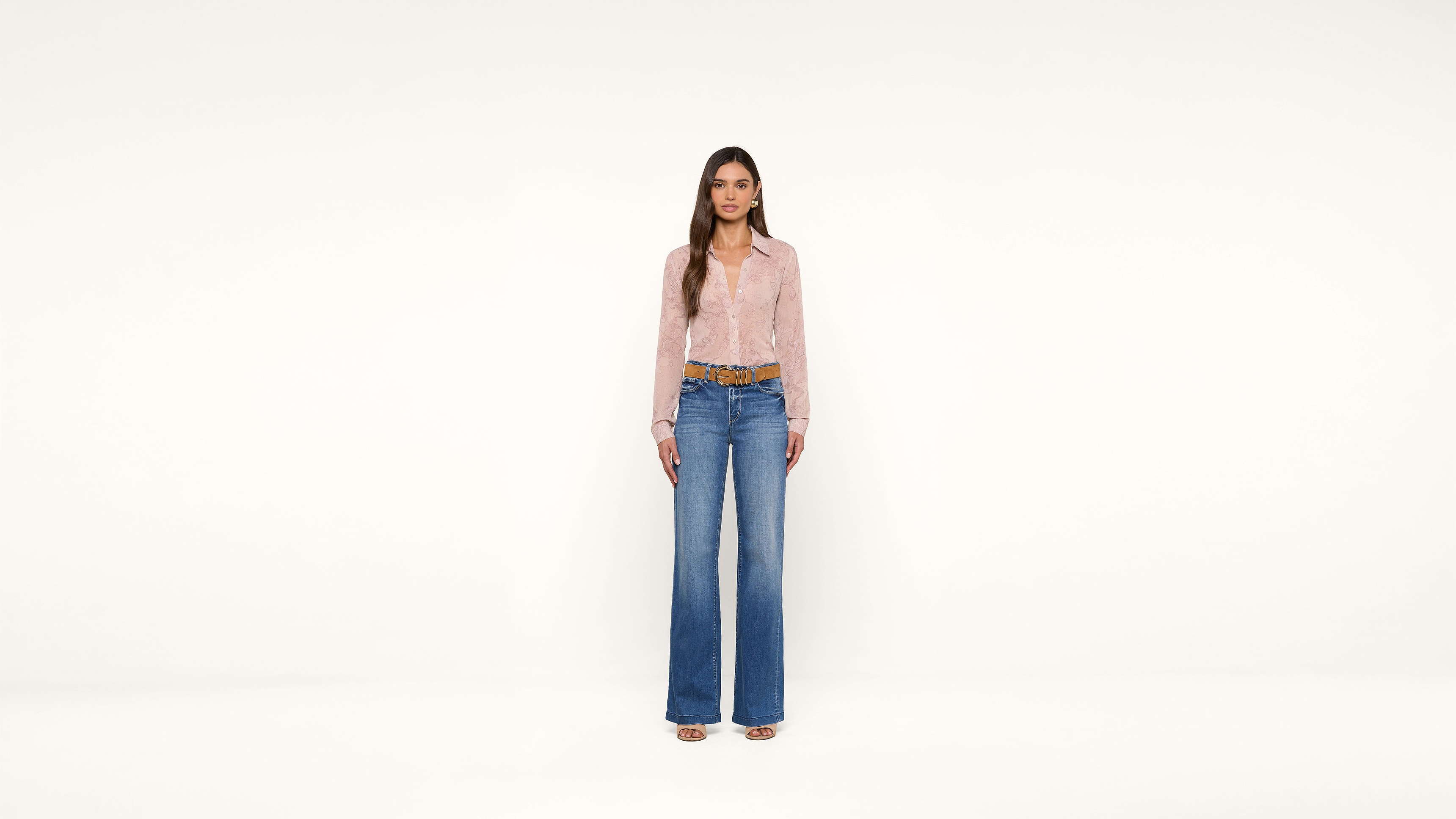 A woman stands against a plain white background, wearing a light pink blouse, wide-leg blue jeans, brown belt, and beige open-toe shoes. She has long brown hair and faces the camera directly.