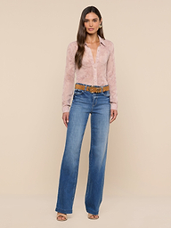 A woman stands against a plain white background, wearing the Laurent Blouse—a light pink, relaxed-fit long sleeve top in sheer poly chiffon—paired with blue high-waisted wide-leg jeans, a brown belt, and open-toed shoes.