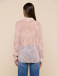 A woman with straight brown hair is shown from the back against a plain background, wearing the Laurent Blouse—a light pink, sheer poly chiffon blouse with a relaxed fit, long sleeves, and a subtle pattern—paired with blue jeans.