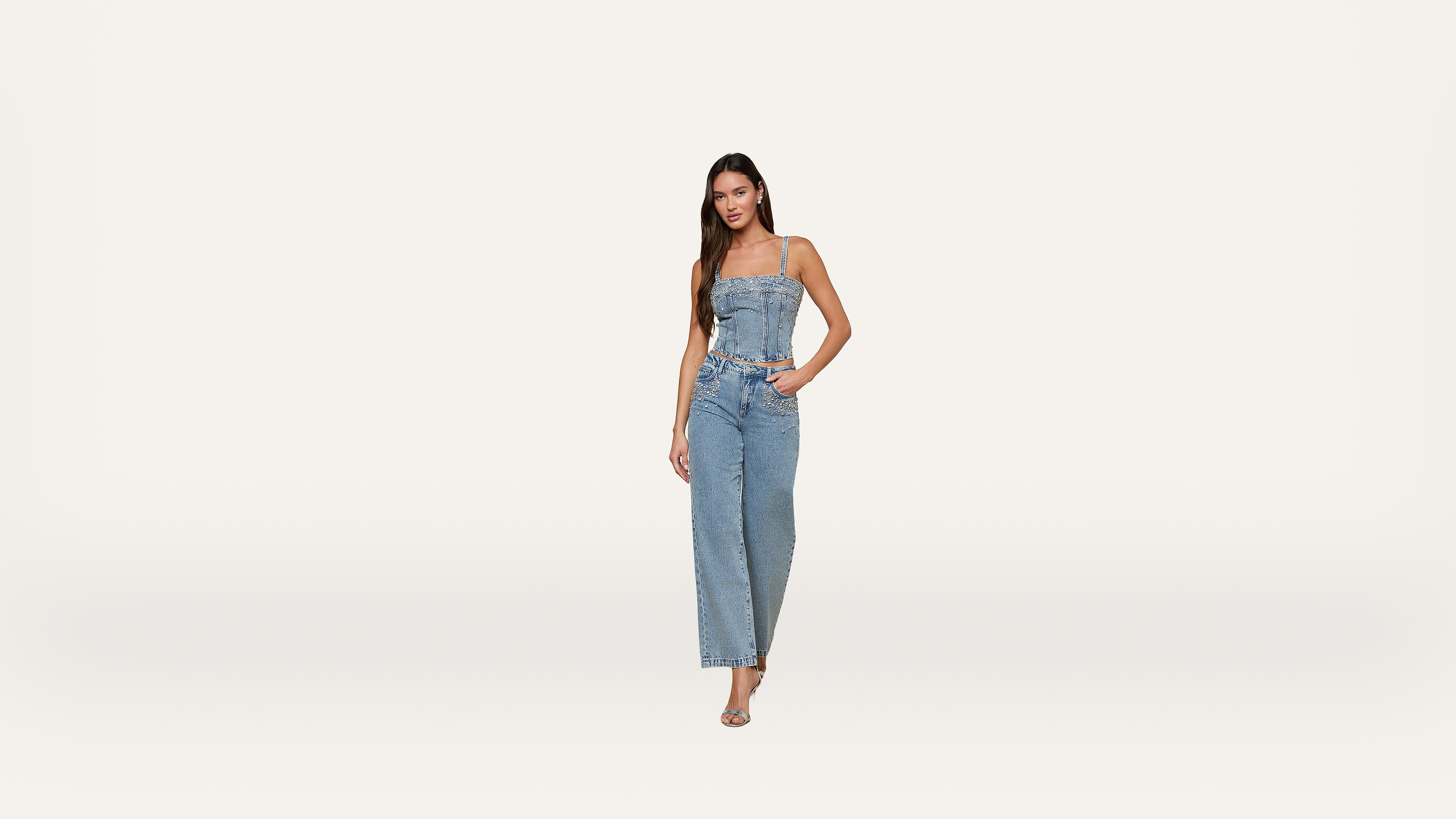 A woman stands against a plain white background, wearing a sleeveless denim jumpsuit and sandals. She has long dark hair and her left hand is in her pocket.