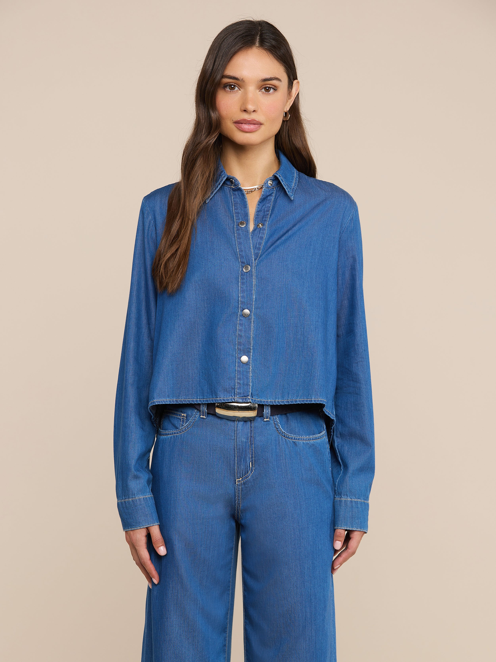 Una mujer con el pelo largo y castaño posa de frente, vestida con la camisa «Annette Chambray» y unos pantalones vaqueros azules a juego, sobre un fondo liso de color beige.