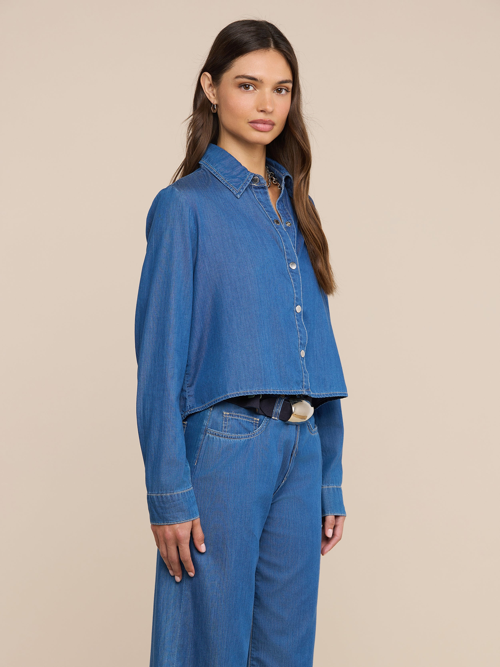 Una mujer con el pelo largo y castaño posa ligeramente de perfil sobre un fondo liso de color beige, con la camisa Annette Chambray —una prenda de corte holgado con bajo asimétrico— combinada con unos vaqueros a juego de pierna ancha.