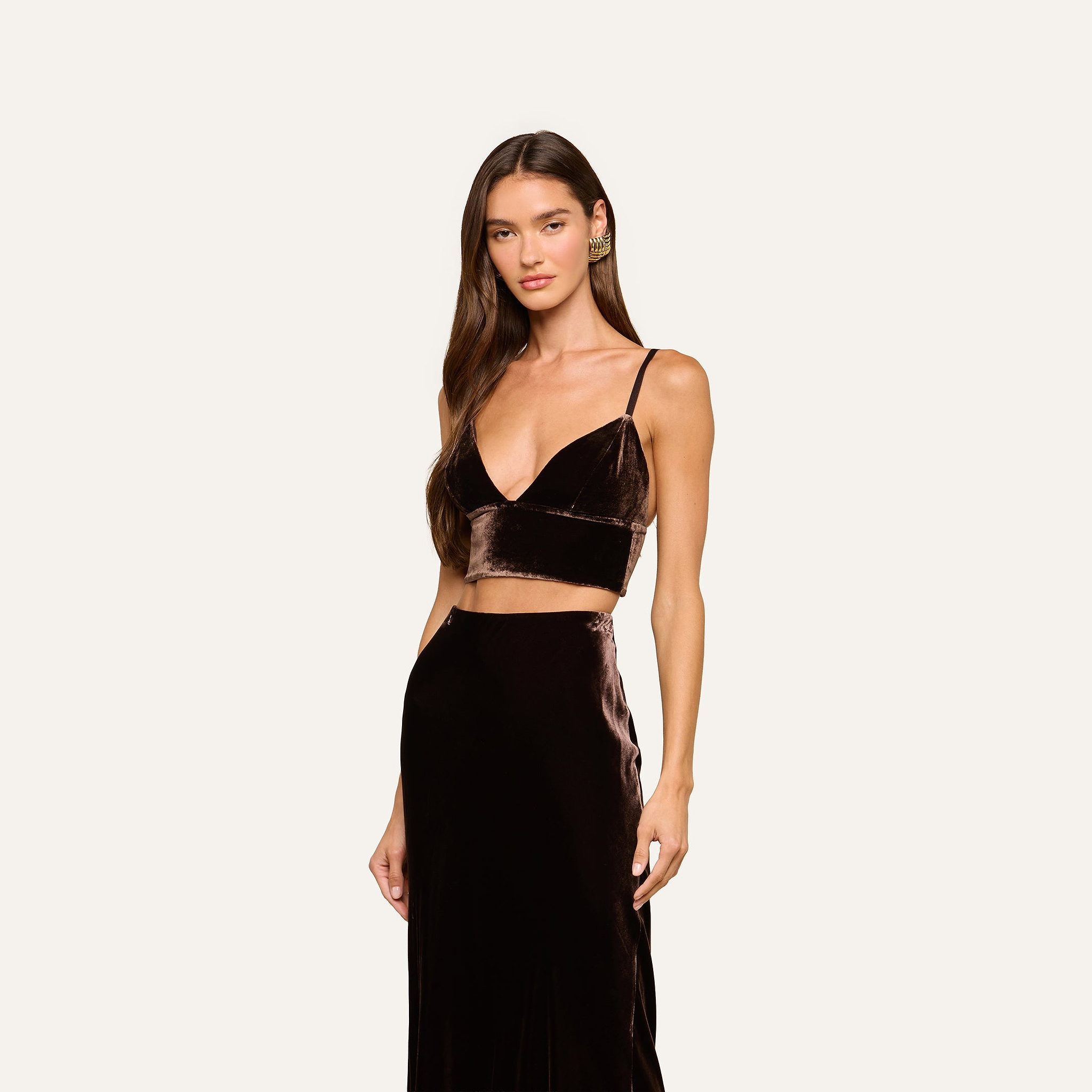 A woman with long brown hair wears the Aislin Velvet Bralette in java brown, featuring adjustable straps and a deep V-neck, paired with a matching high-waisted skirt, standing against a plain light background.