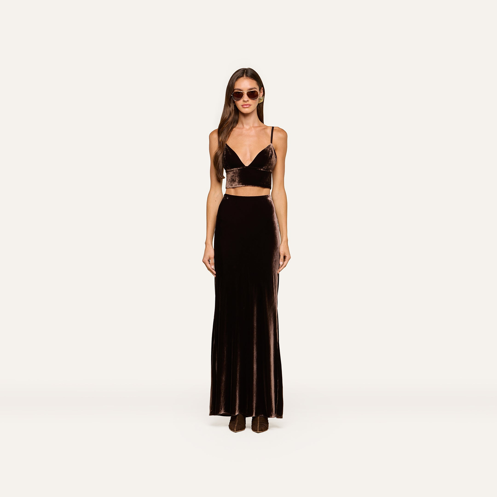 Woman wearing sunglasses, an Aislin Velvet Bralette in java brown with adjustable straps, and a matching long velvet skirt, standing against a plain white background.