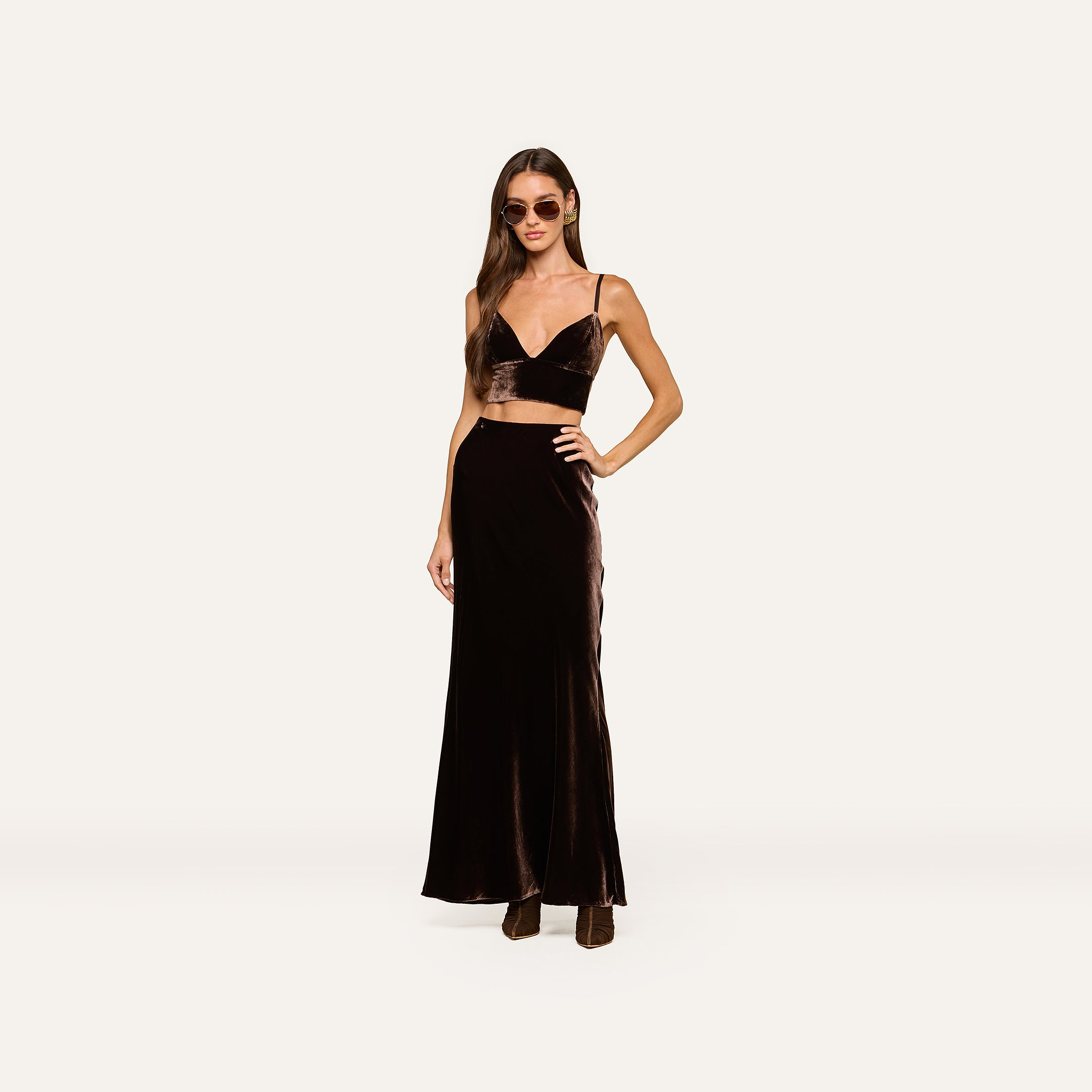 A woman wearing sunglasses, the Aislin Velvet Bralette in java brown with adjustable straps, a matching long skirt, and boots stands against a plain light background.