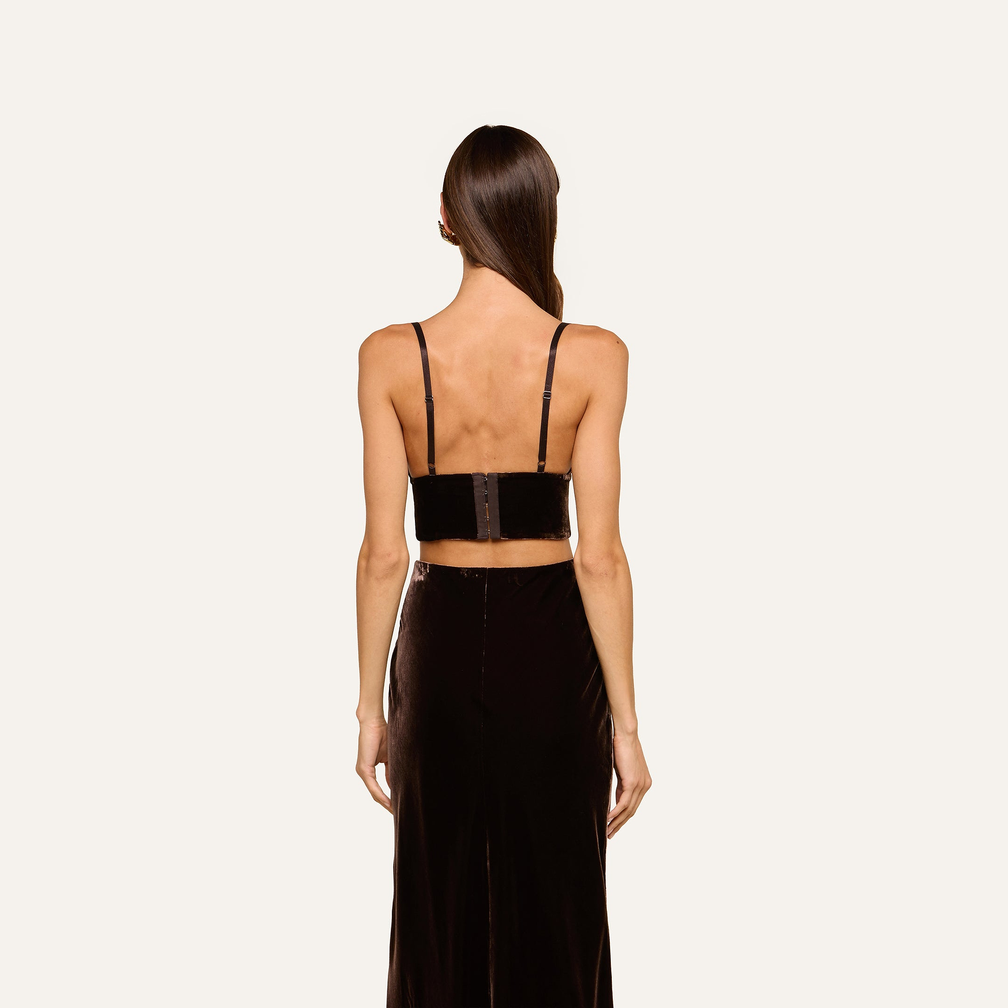 A woman with long straight hair is shown from the back against a plain light background, wearing the Aislin Velvet Bralette—a black velvet crop top with thin, adjustable straps—paired with a matching high-waisted skirt.