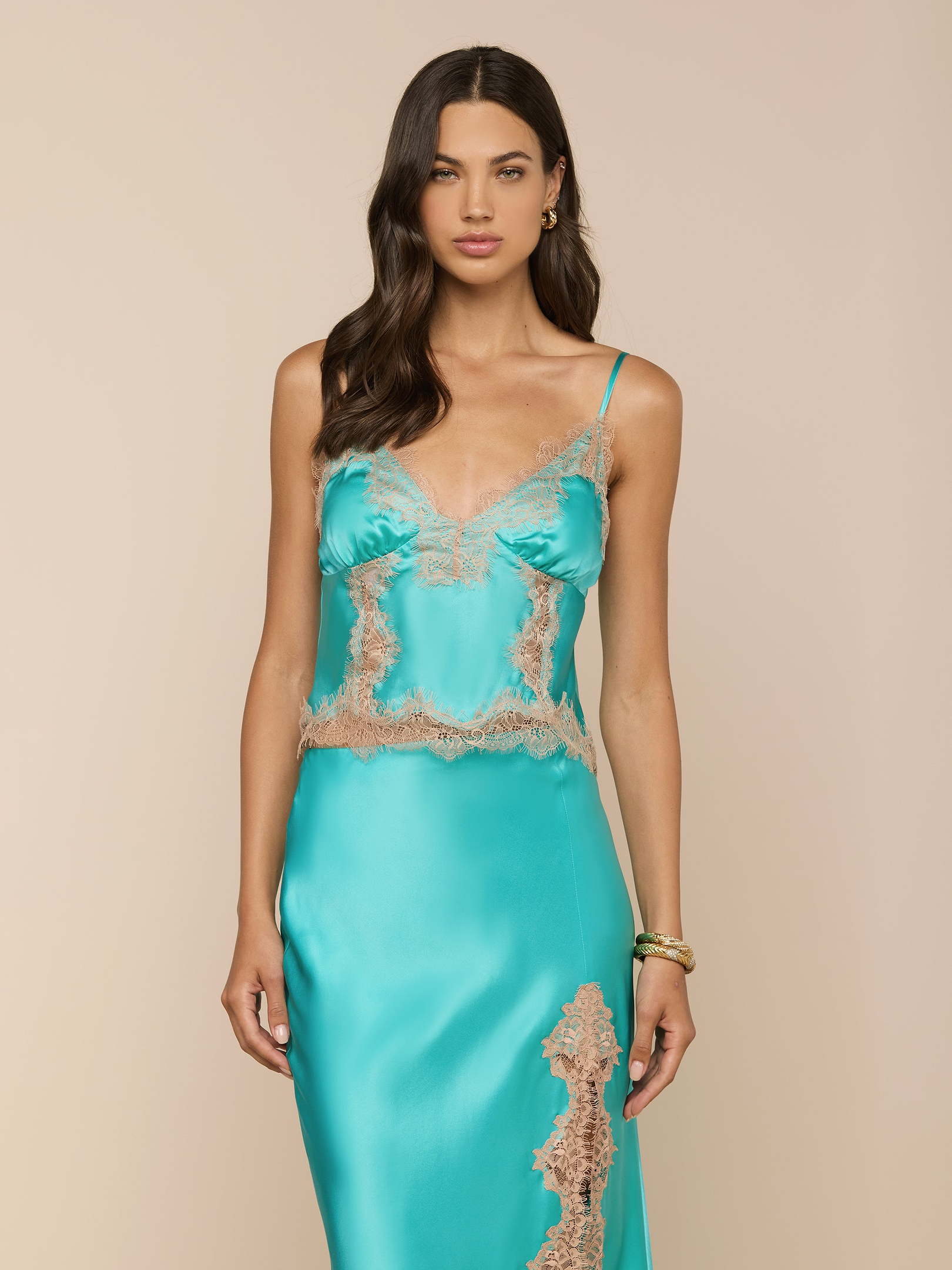 Woman wearing the Kimber Silk Lace Camisole in turquoise, featuring delicate lace trim, standing against a plain light background. She has long, wavy dark hair and accessorizes with gold hoop earrings and a bracelet.