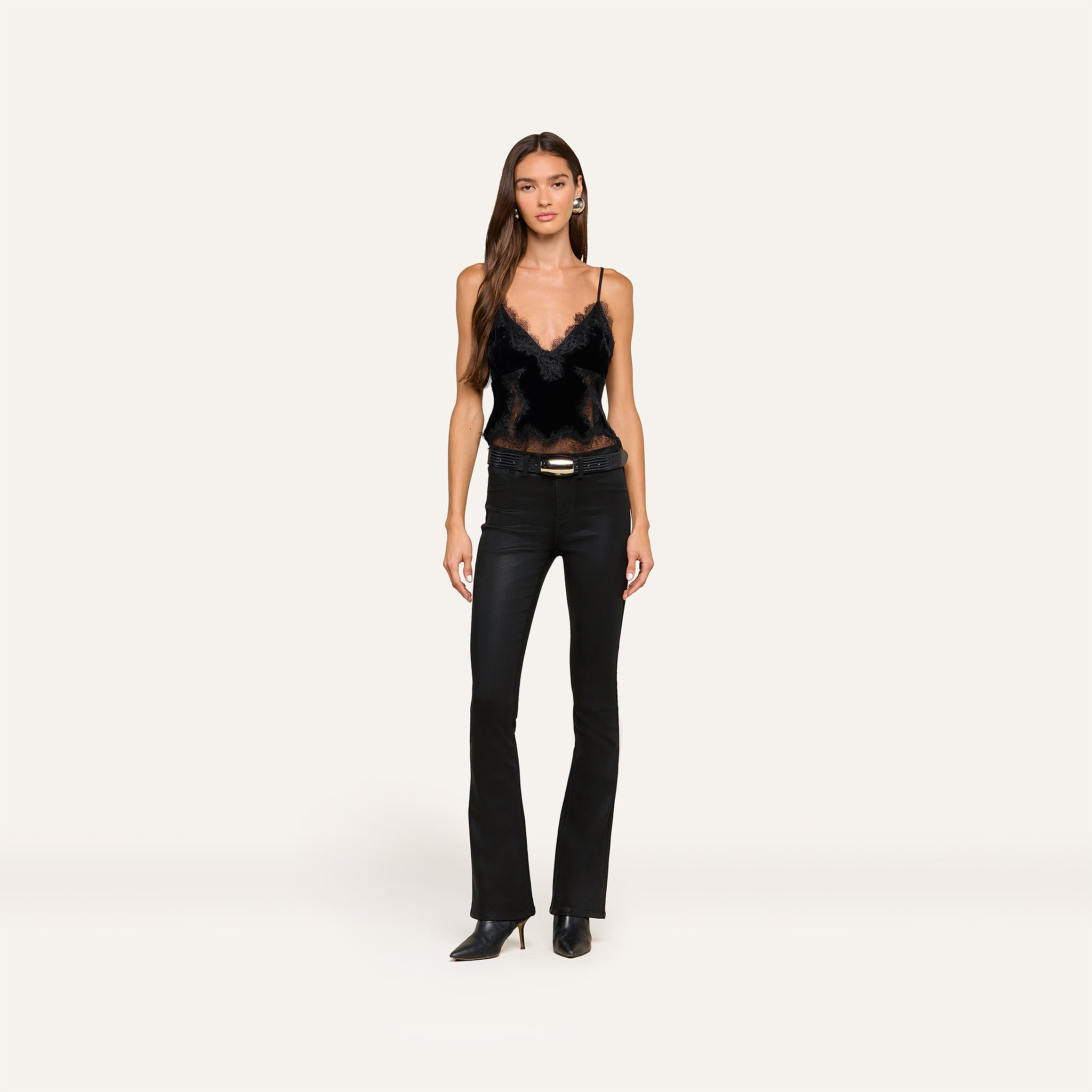 A woman with long brown hair and a neutral expression stands against a plain background, wearing the Kimber Velvet Lace Camisole, paired with black fitted jeans, a belt, and black pointed-toe boots.