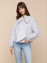 A woman wears the Deon Tie-Neck Blouse—an oversized blue and white vertical striped blouse with blouson sleeves—paired with light blue jeans, standing against a plain light background.