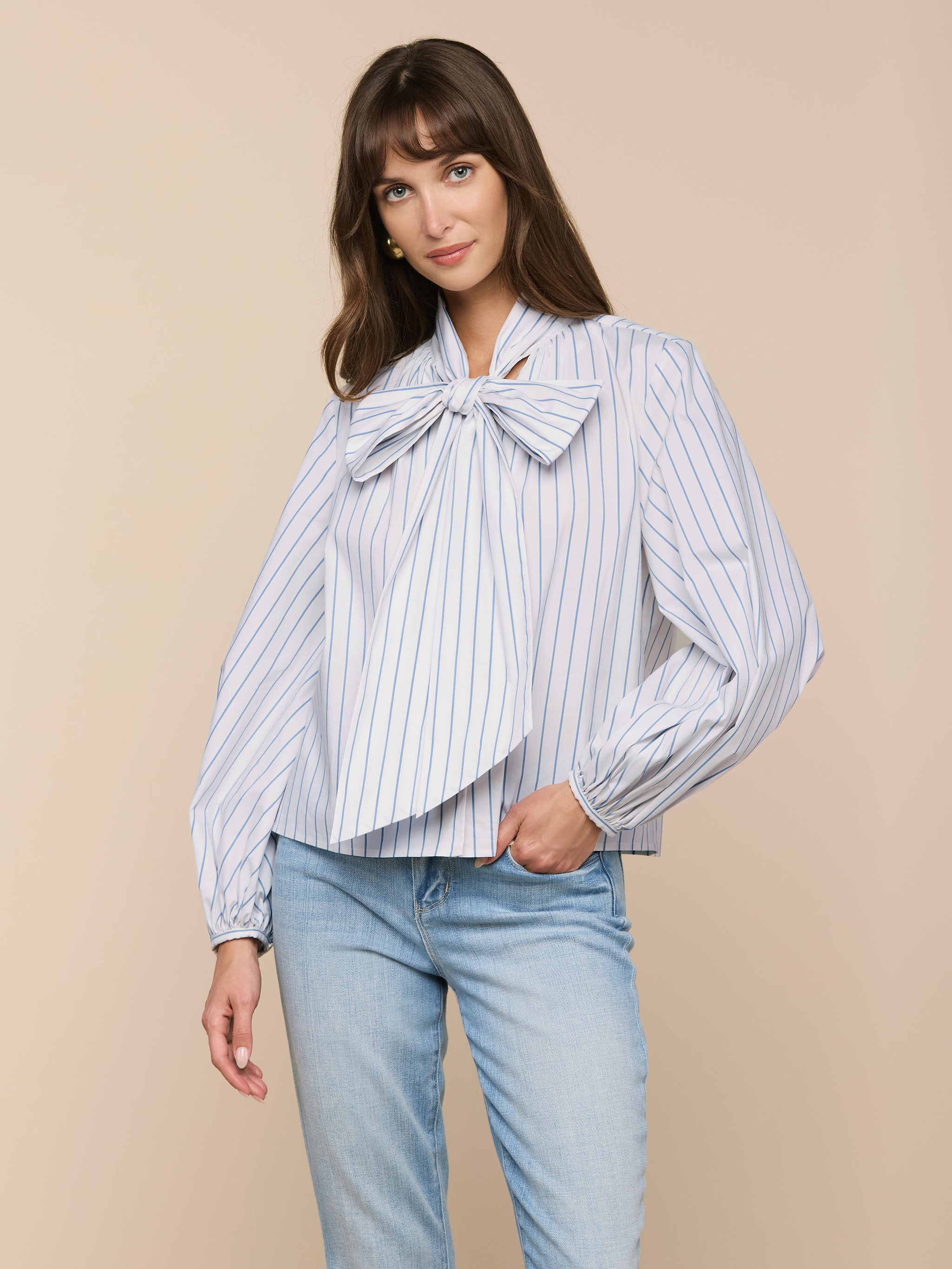 A woman wears the Deon Tie-Neck Blouse—an oversized blue and white vertical striped blouse with blouson sleeves—paired with light blue jeans, standing against a plain light background.
