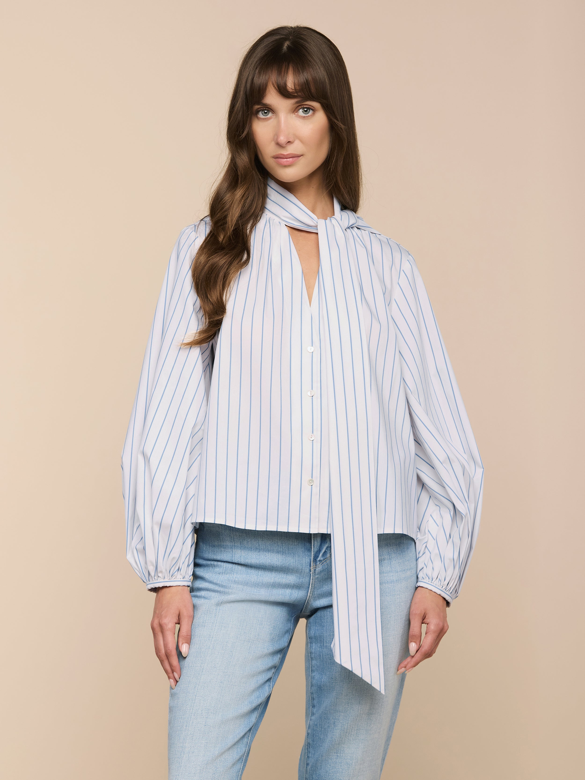 A woman wearing the Deon Tie-Neck Blouse—an oversized blouse crafted from white and blue vertical stripe organic cotton-blend fabric, designed with long blouson sleeves and a long scarf tie at the neck—paired with light blue jeans, stands against a plain light background.