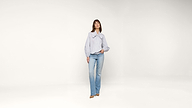A woman stands against a plain white background, wearing a light blue and white striped blouse with a large bow and light blue jeans. She has one hand in her pocket and is looking forward.
