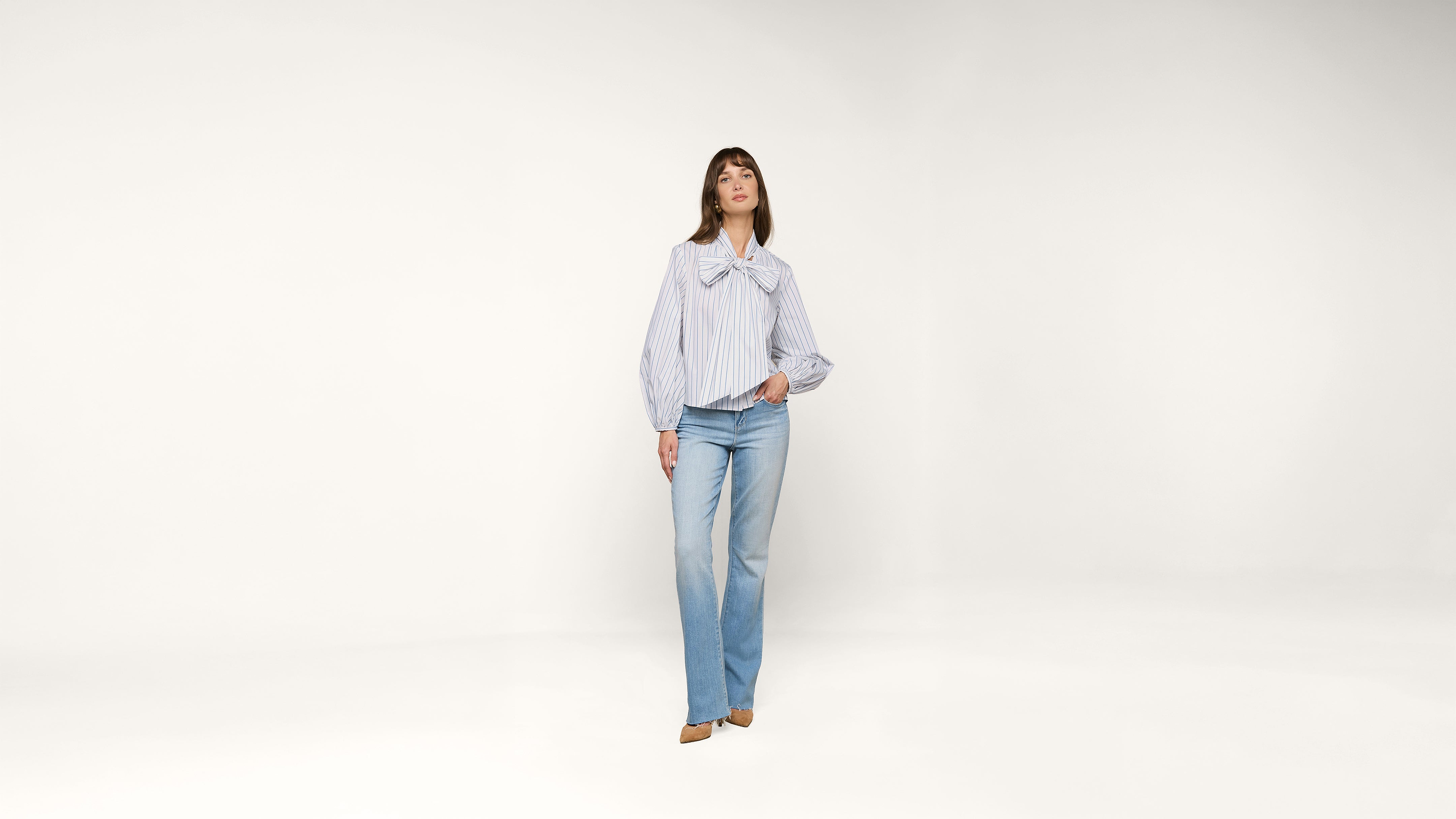 A woman stands against a plain white background, wearing a light blue and white striped blouse with a large bow and light blue jeans. She has one hand in her pocket and is looking forward.