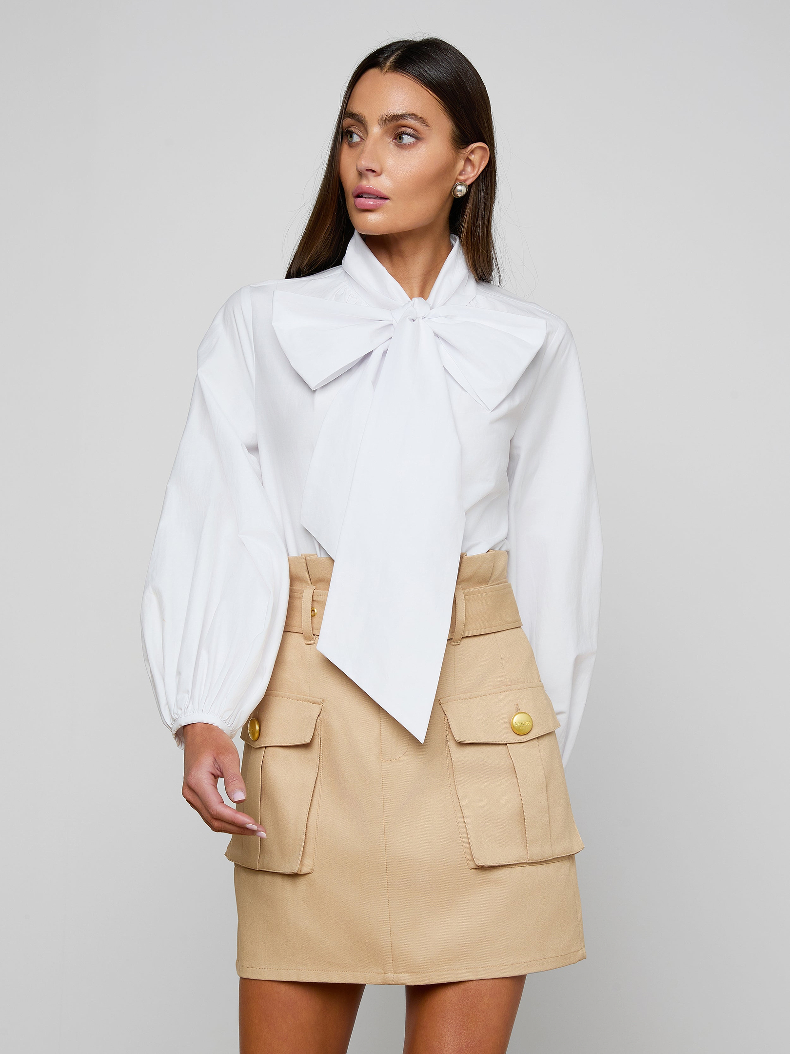 A woman stands against a plain background, wearing the Deon Tie-Neck Blouse—an organic cotton poplin top with blouson sleeves and an oversized neck tie—paired with a beige skirt featuring front pockets and gold buttons. Her long, straight hair frames her face as she gazes slightly to the side.