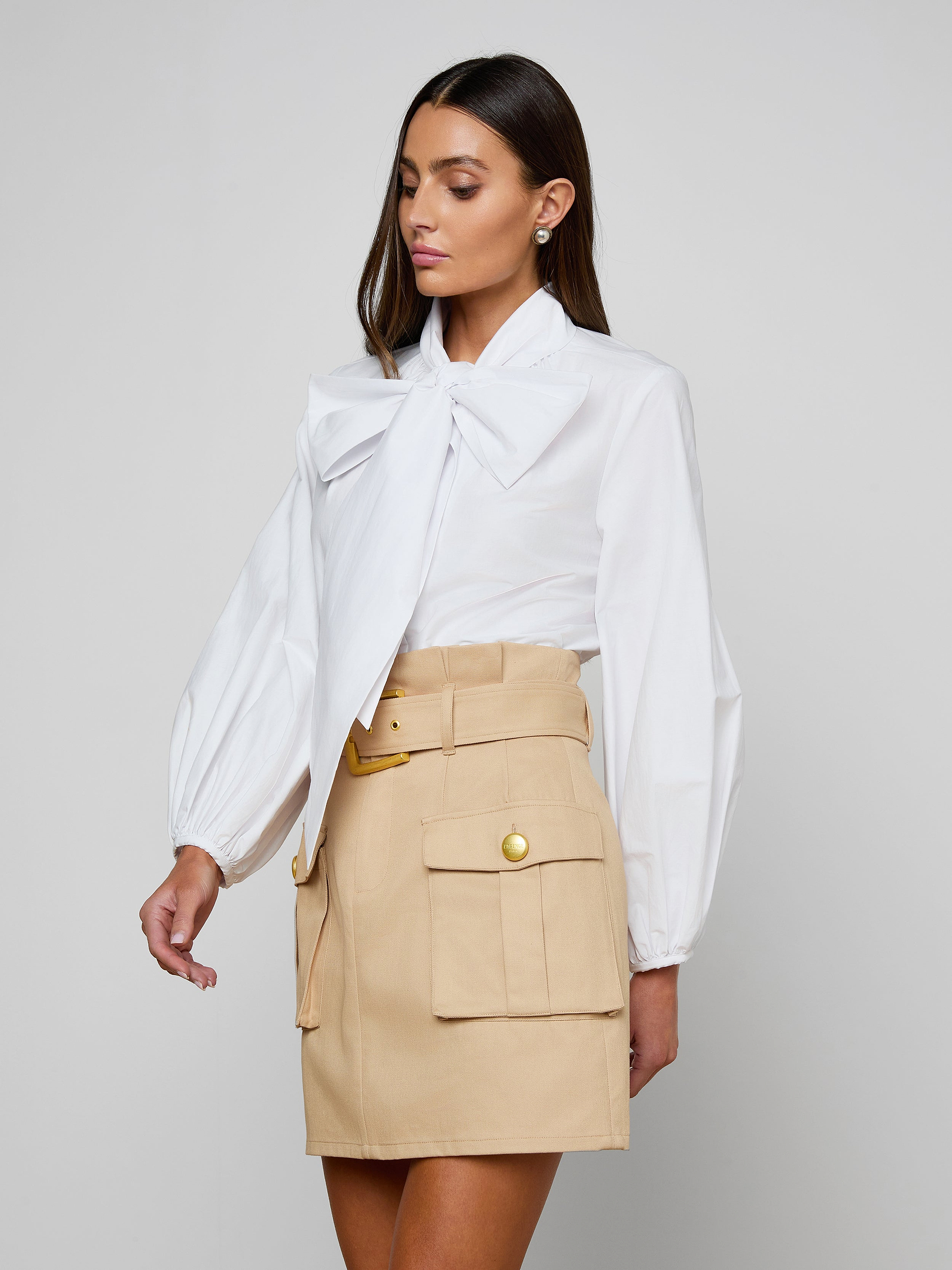 A woman is wearing the Deon Tie-Neck Blouse—a white organic cotton poplin top with blouson sleeves and an oversized neck tie—paired with a beige high-waisted skirt featuring front pockets and gold buttons, standing against an off-white background.