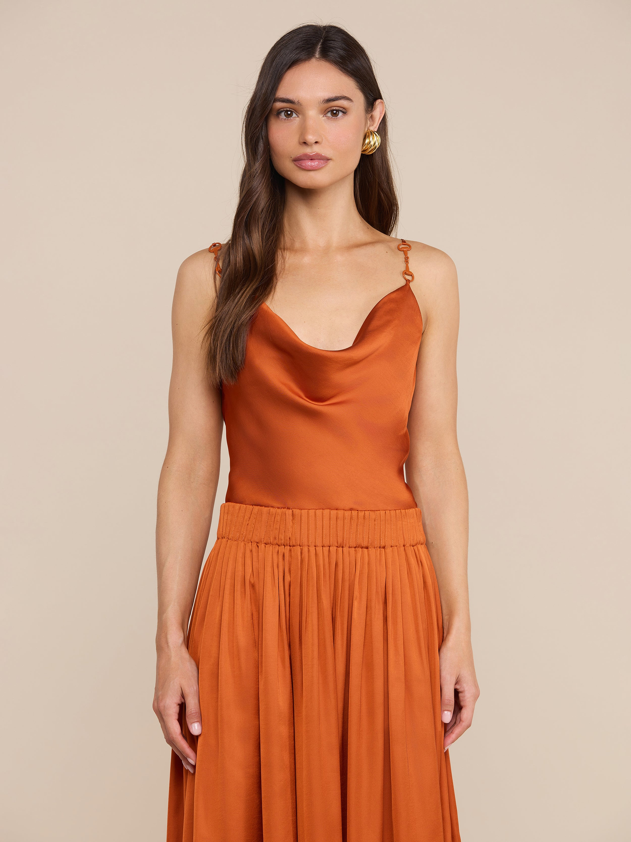 A woman wearing the Calista Satin Camisole in burnt orange, paired with a matching pleated skirt, stands against a plain light background.