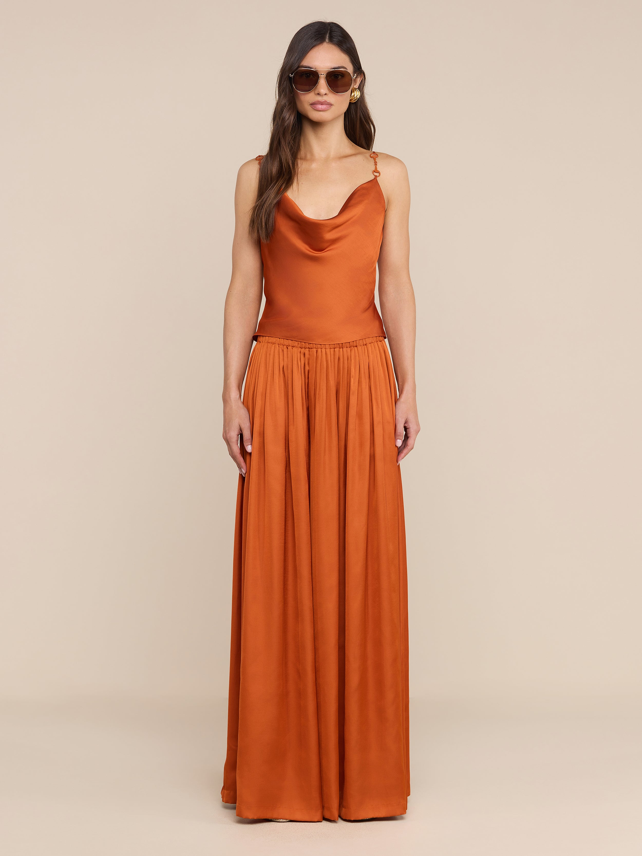 A woman wearing sunglasses and an orange, sleeveless, floor-length dress stands facing forward against a plain white background. The outfit is styled with the Calista Satin Camisole, adding a stylish touch to the overall look.