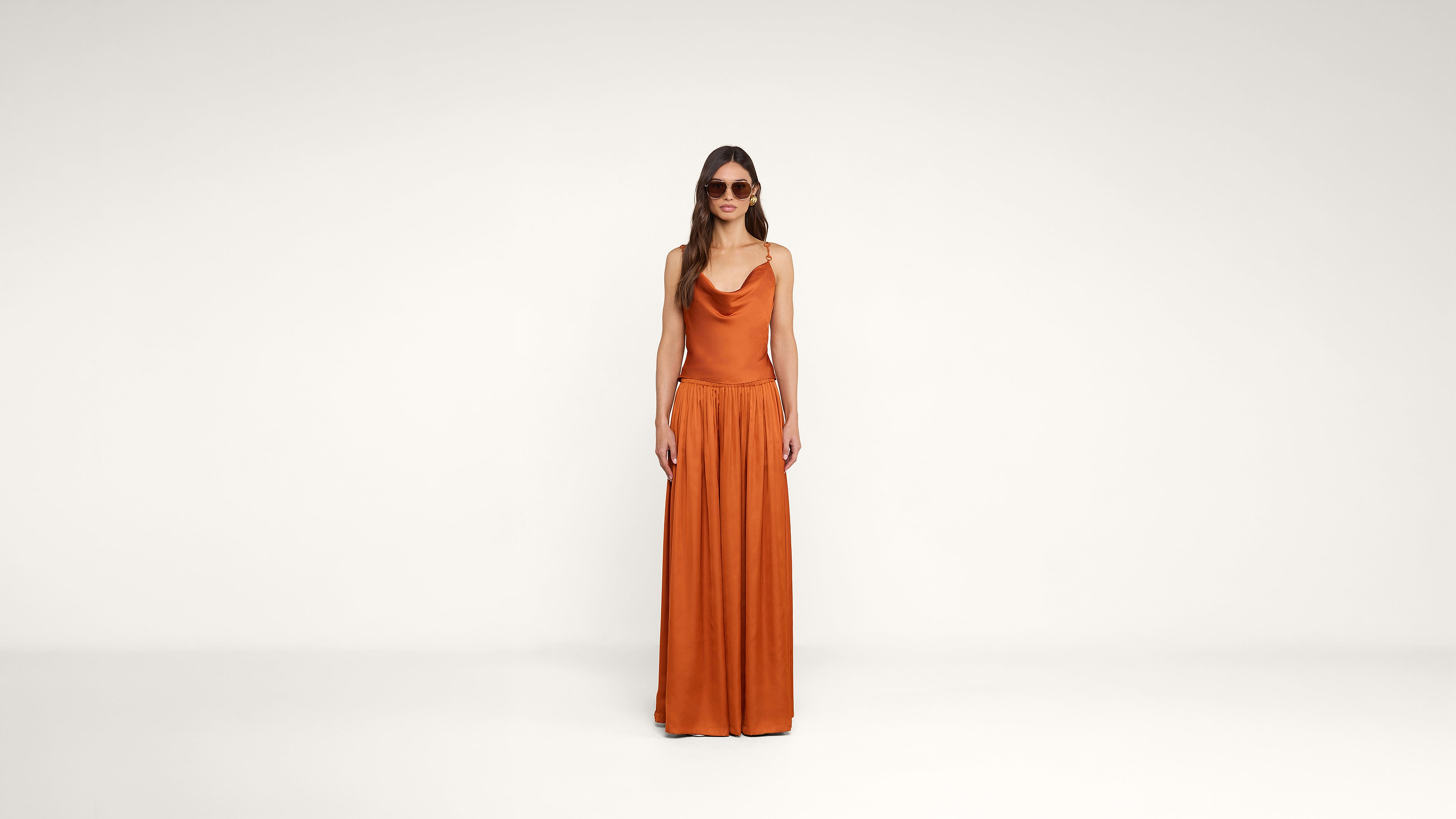 A woman stands against a plain white background, wearing sunglasses and an orange sleeveless dress with a loose skirt.