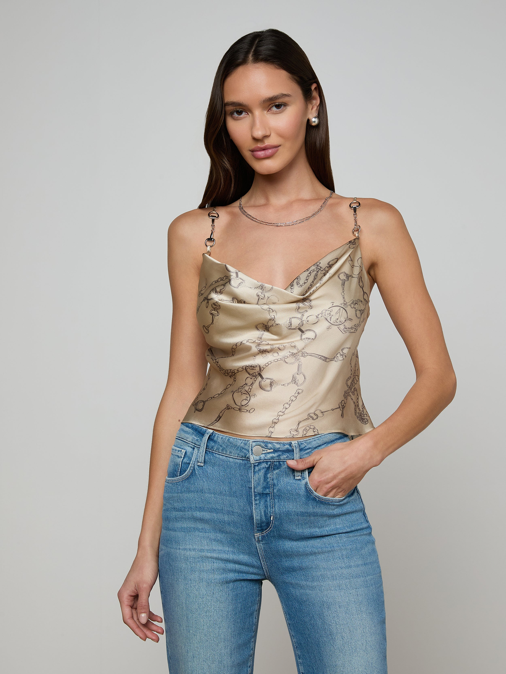 A woman with long dark hair wears the Calista Silk Camisole, a cropped top featuring a horse bit chain print, paired with blue jeans and standing against a plain light background.