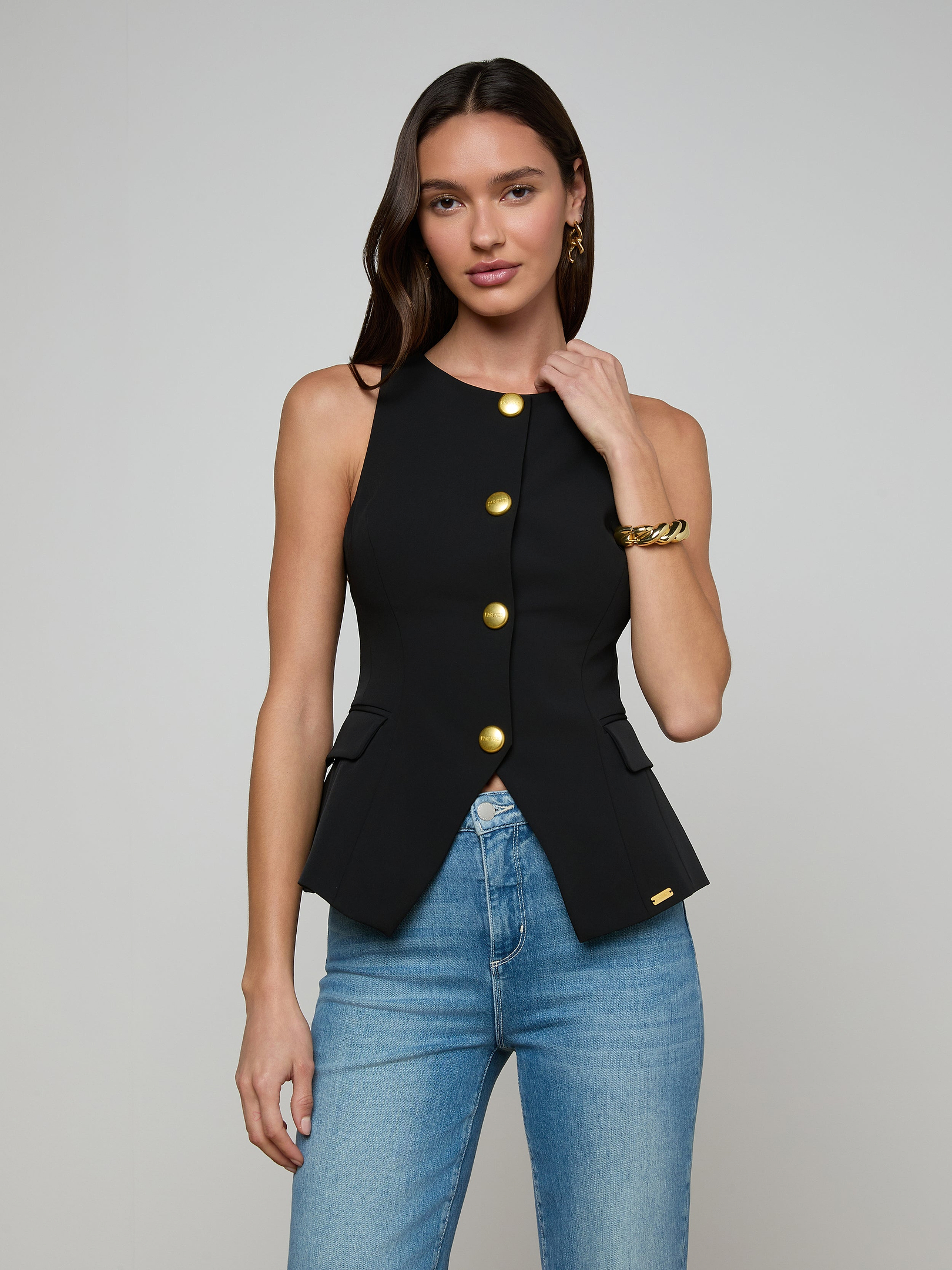 A woman wearing the Tessa Vest, a sleeveless black top with gold buttons, paired with blue jeans stands against a plain light background, touching her hair.