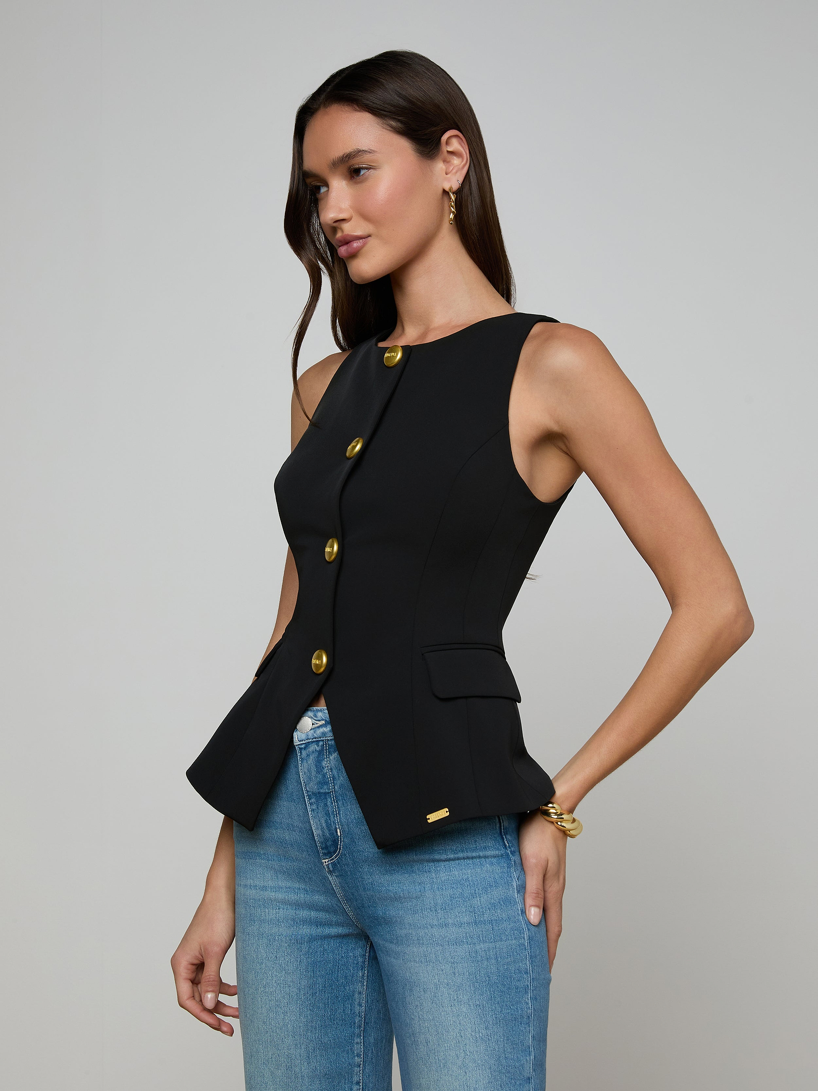 A woman wearing the Tessa Vest—a sleeveless suit vest with gold buttons—paired with blue jeans stands against a plain light background.