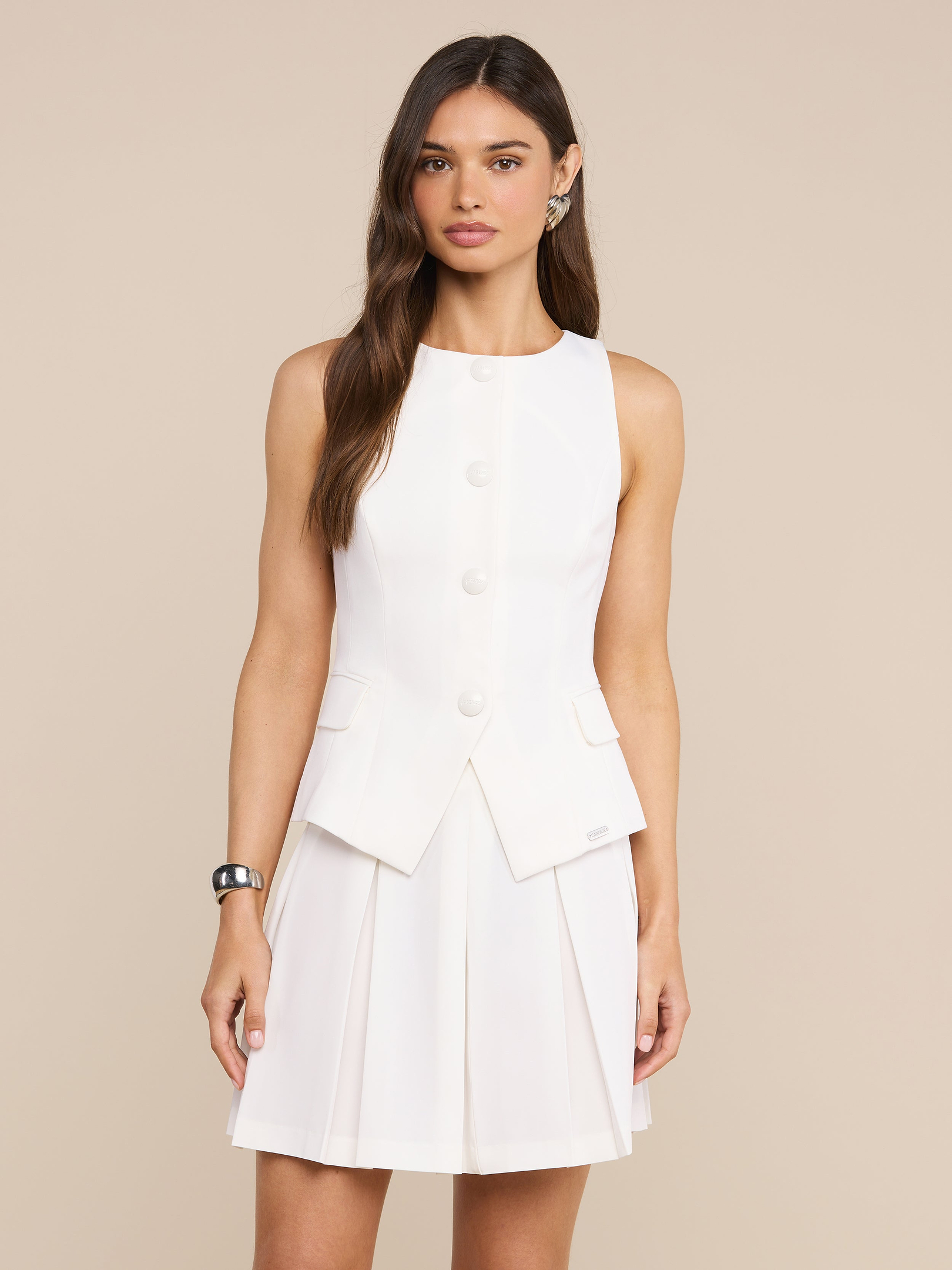 Una donna con lunghi capelli castani indossa il Gilet Tessa bianco senza maniche, con grandi bottoni e gonna a pieghe in stile blazer minimalista. Si staglia su uno sfondo beige, accessoriato con un braccialetto nero e orecchini dargento.