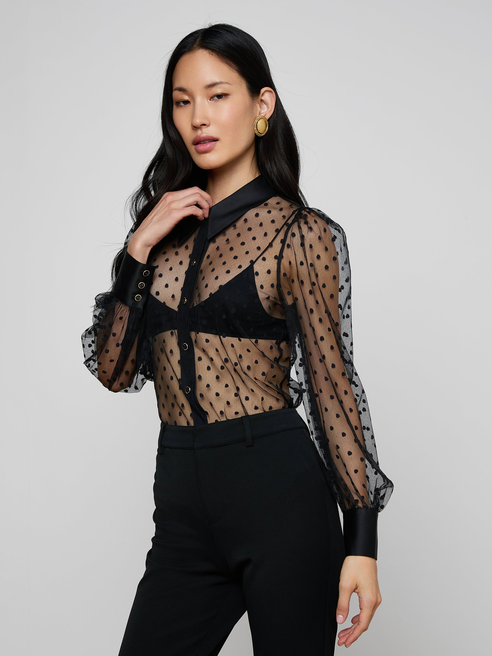 Wearing the Sophie Long-Sleeved Blouse—a sheer Swiss dot top with black cuffs and collar—layered over a black bra and paired with high-waisted black pants, she poses against a plain light background.