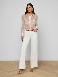 A woman with long hair and a neutral expression stands against a white background, wearing the Sophie Long-Sleeved Blouse—a sheer Swiss dot top—paired with white wide-leg pants, a gold belt, gold earrings, and open-toe heels.