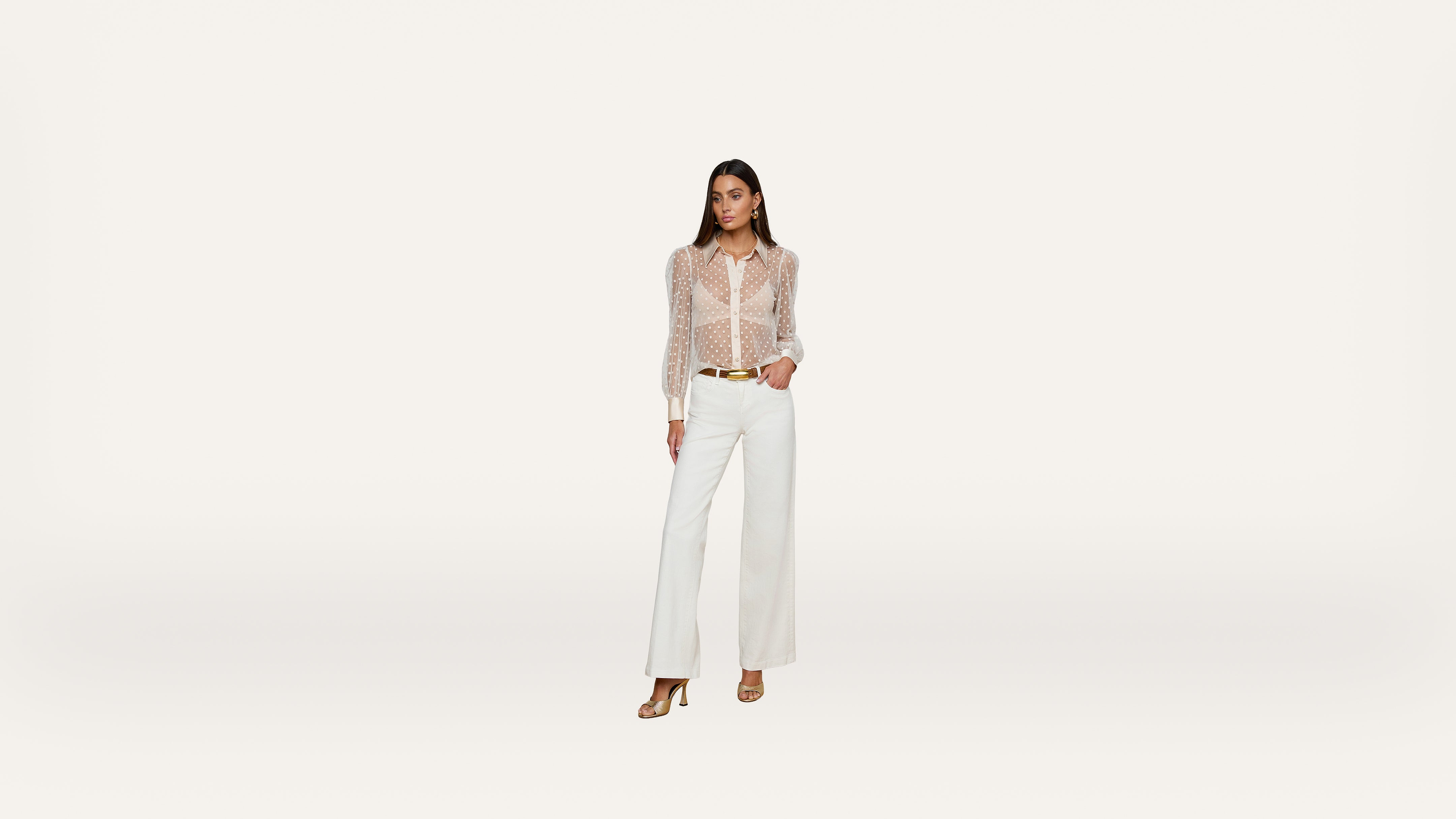 A woman stands confidently in a sheer, polka dot blouse and white wide-leg pants with heels, against a plain, light background.