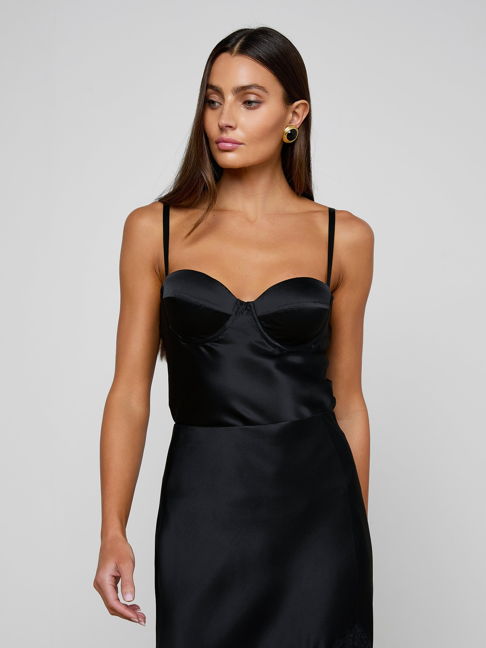 A woman with long brown hair is wearing the Orlina Silk Camisole—a black satin sleeveless top with thin straps and underwire cups—standing against a plain light background and looking to her left.
