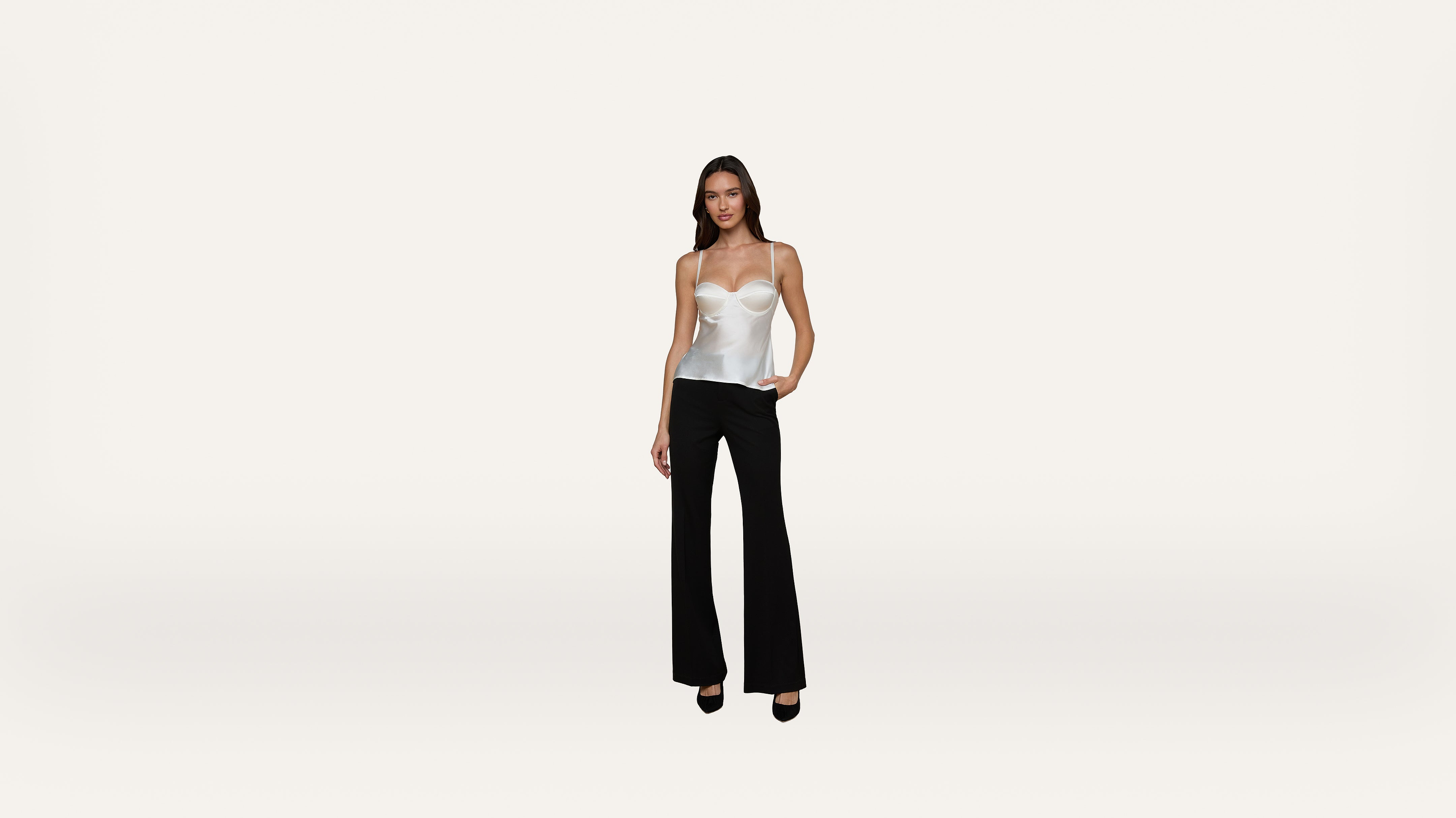 A woman stands against a plain white background, wearing a white spaghetti strap top, black flared pants, and black heels, with one hand in her pocket.