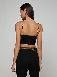 Woman with long brown hair wearing the Sophia Chain Bralette, a black crop top with gold chain straps and structured silhouette, paired with black high-waisted pants made from stretch suiting fabric, facing away from the camera against a plain light background.
