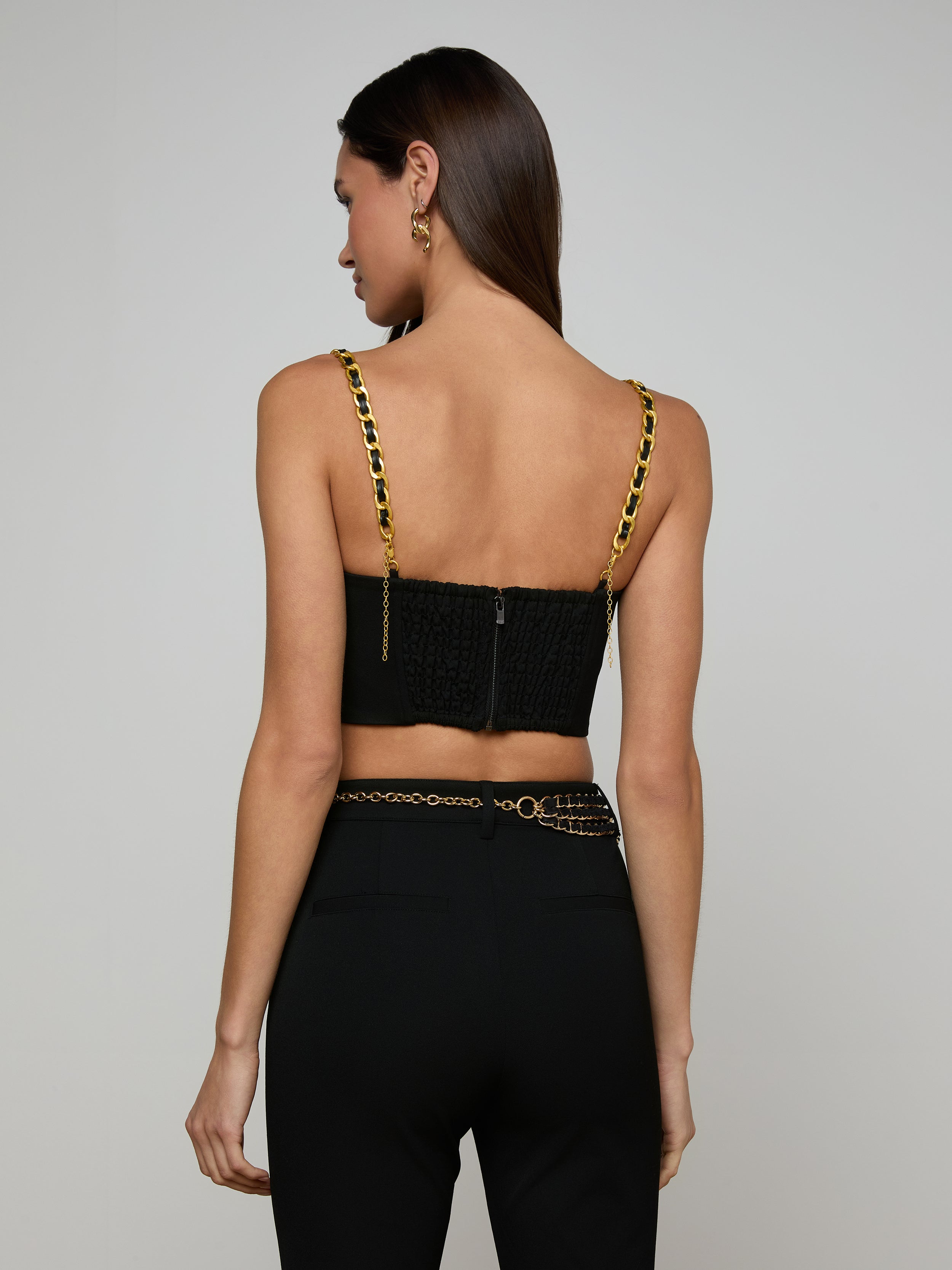 Woman with long brown hair wearing the Sophia Chain Bralette, a black crop top with gold chain straps and structured silhouette, paired with black high-waisted pants made from stretch suiting fabric, facing away from the camera against a plain light background.