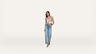 A woman stands against a plain white background, wearing a floral tank top, wide-leg blue jeans, and beige heels. She has one hand in her pocket and is smiling slightly.