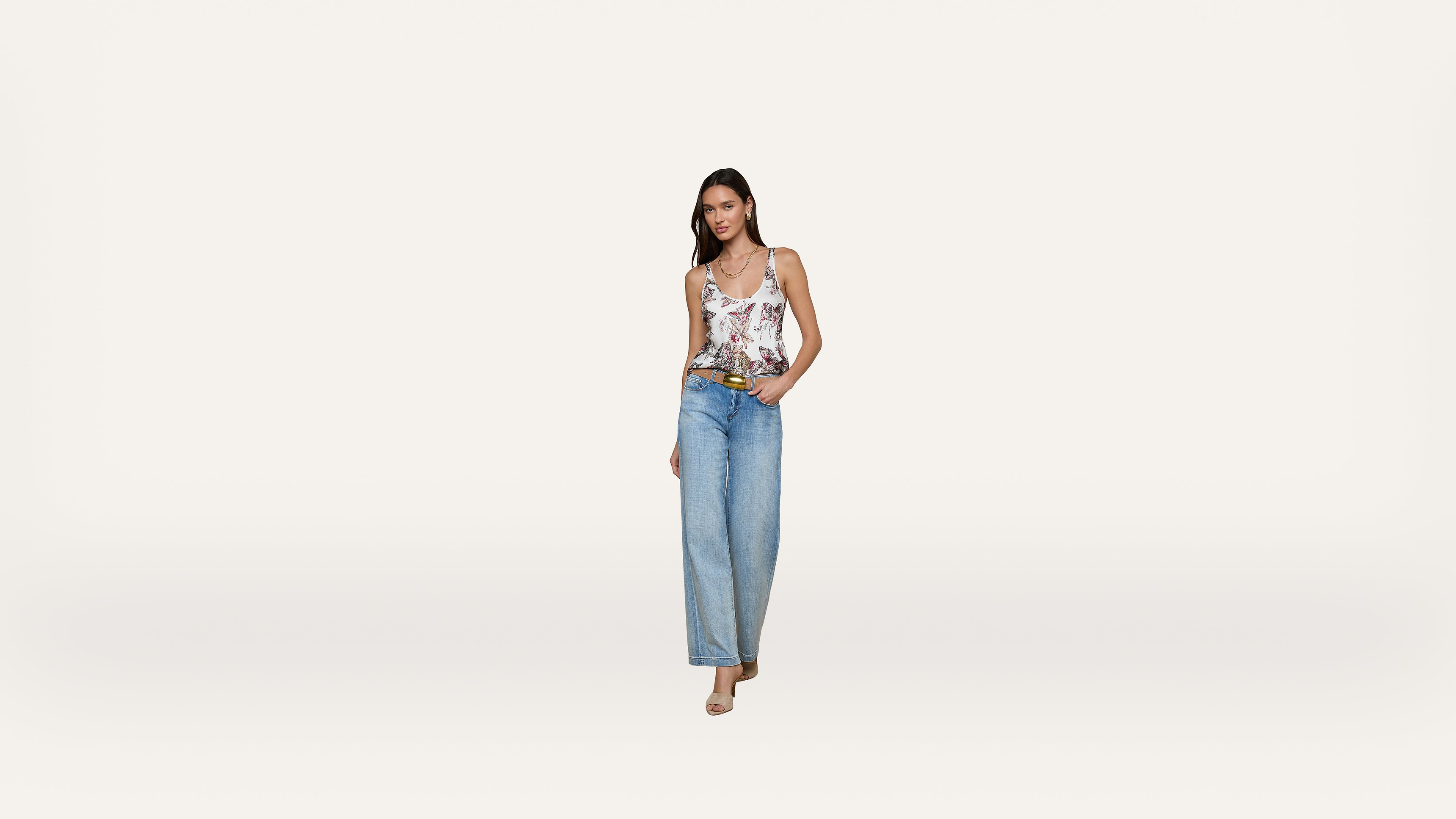 A woman stands against a plain white background, wearing a floral tank top, wide-leg blue jeans, and beige heels. She has one hand in her pocket and is smiling slightly.
