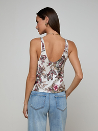 A woman with long brown hair, wearing the Eddie Tank—featuring a vibrant floral print—and light blue jeans, stands with her back to the camera against a plain light background.