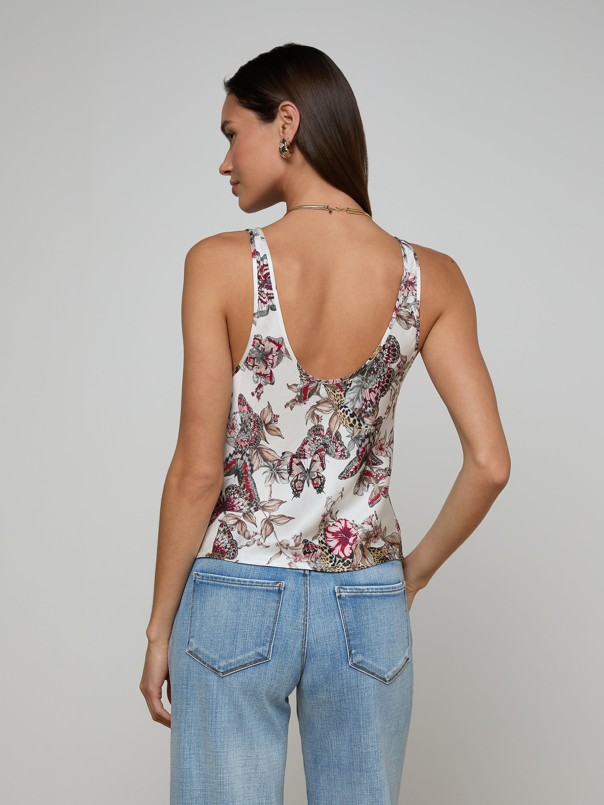 A woman with long brown hair, wearing the Eddie Tank—featuring a vibrant floral print—and light blue jeans, stands with her back to the camera against a plain light background.