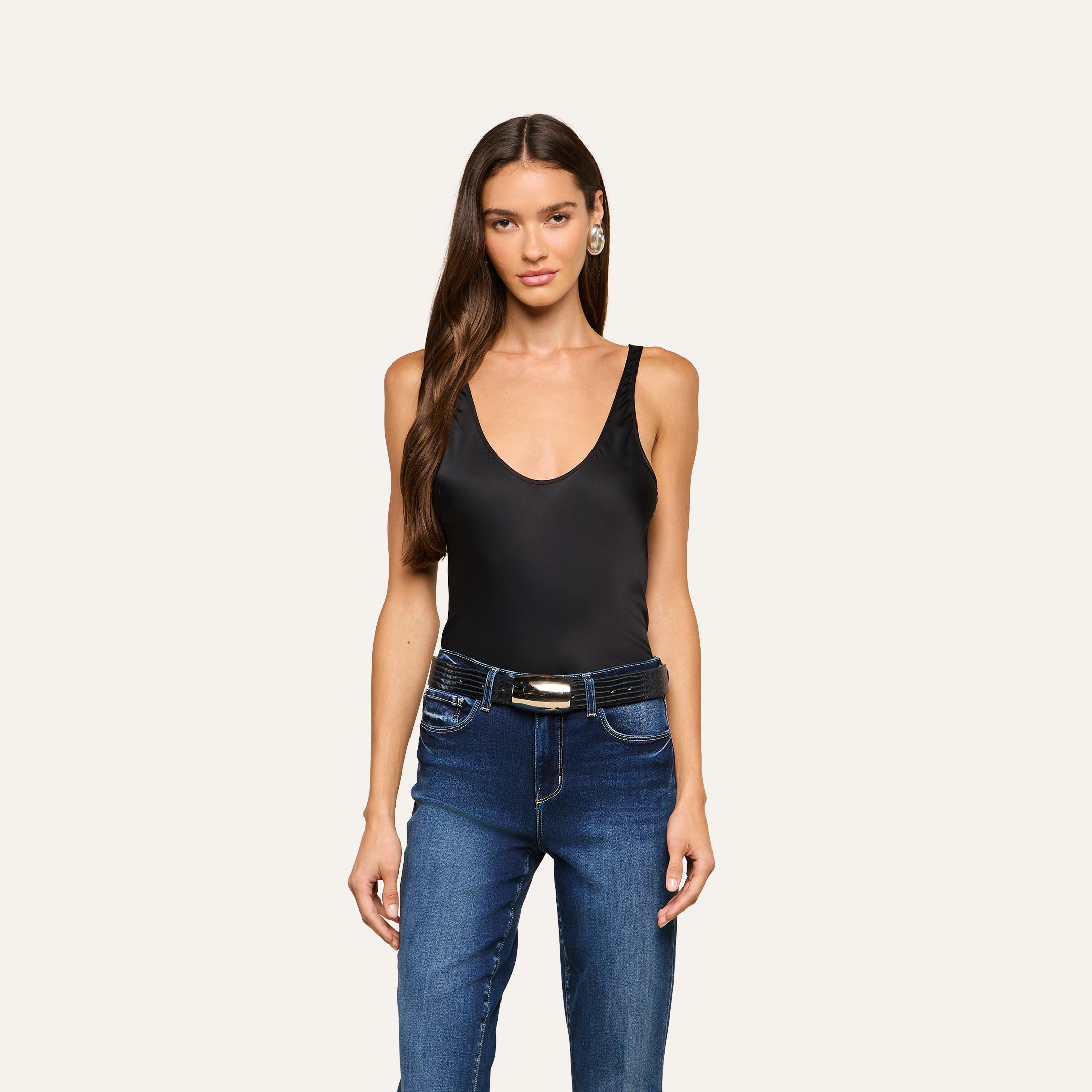A woman with long brown hair is wearing the Eddie Tank—a sleeveless black bodysuit with a flattering drape—paired with blue jeans and a belt, standing against a plain light background.