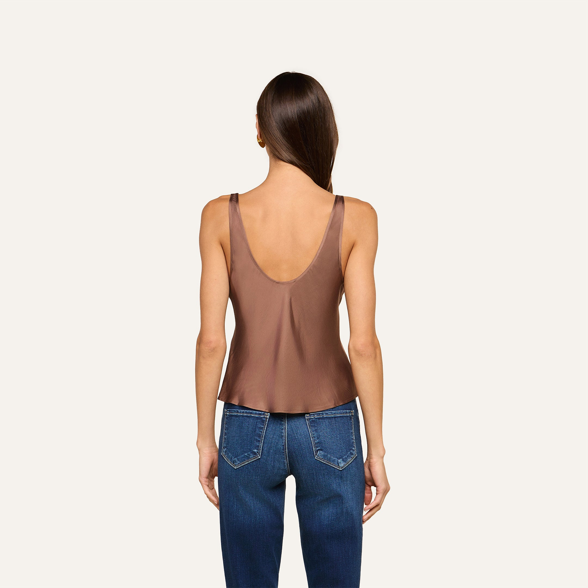 A woman with long brown hair stands facing away, wearing the Eddie Tank in cardamom brown—a sleeveless top with a low scoop neckline—paired with blue jeans against a plain off-white background.