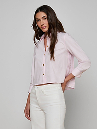 A woman with long brown hair wears the Fawn Shirt—a cropped pale pink top—paired with high-waisted white pants, standing against an off-white background and looking at the camera.